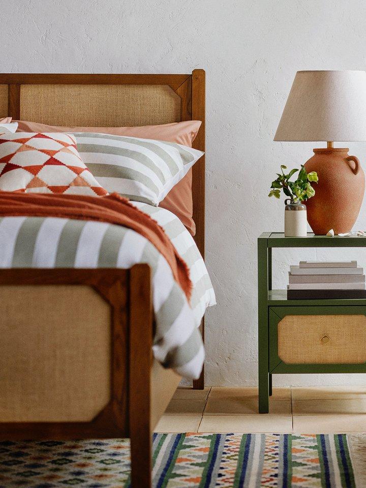 A bedroom scene showing a bed, a side table with a lamp, and a potted plant