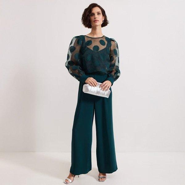 Woman in sheer polka dot top and teal trousers holding a silver clutch.