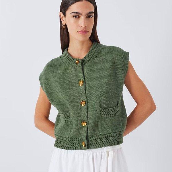 A woman wears a short-sleeved, olive green knit cardigan with gold buttons and front pockets, paired with a white skirt.