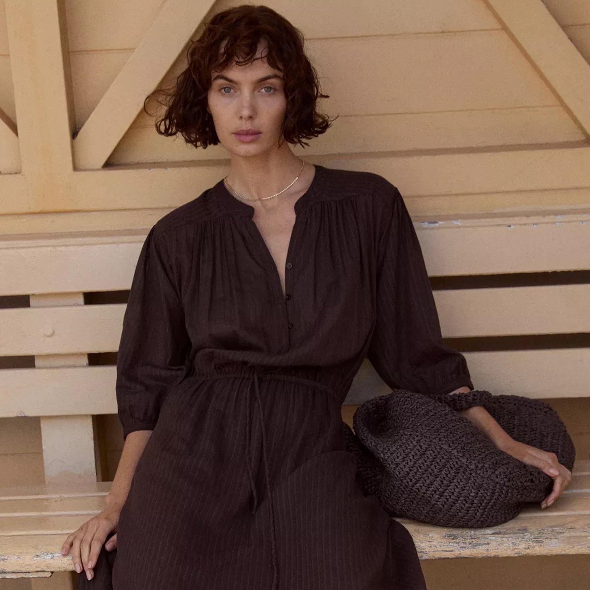 Woman in a dark brown, flowy dress with a cinched waist, sitting on a bench with a matching knitted bag.