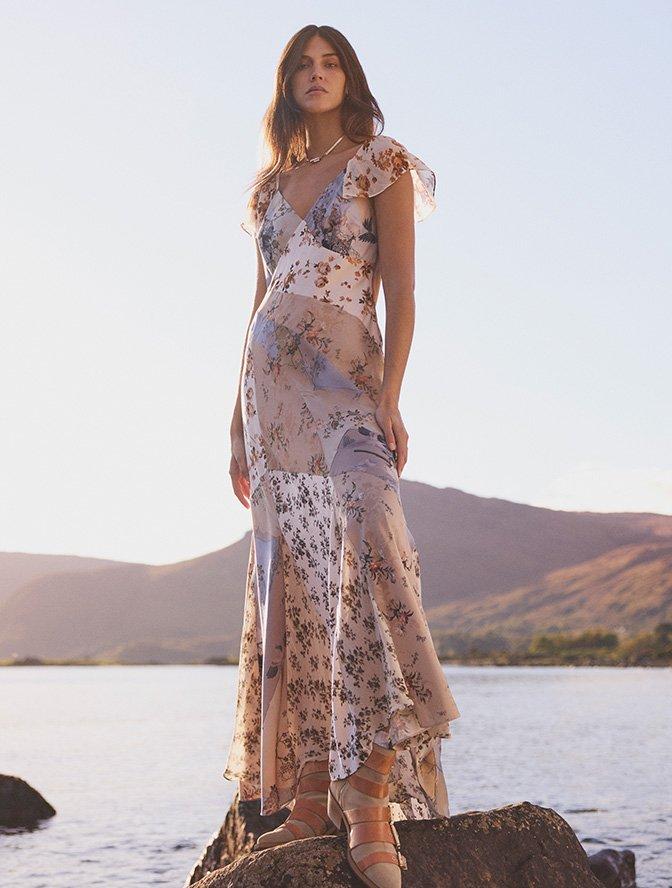A woman in a floral maxi dress standing on a rock by the water with mountains in the background.