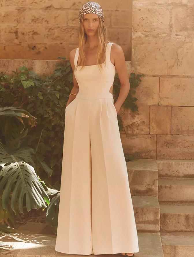 A woman in a white sleeveless jumpsuit with a patterned headscarf, standing against a stone wall with greenery.