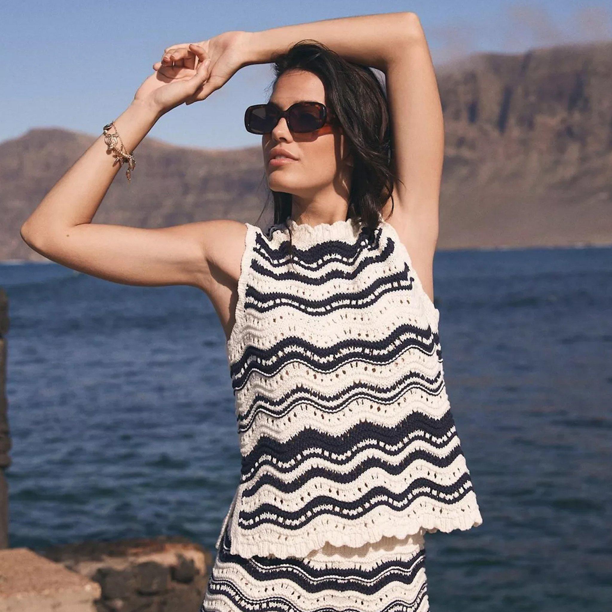 A woman wearing sunglasses and a sleeveless, wavy black-and-white knitted top, posing outdoors near water with mountains in the background.