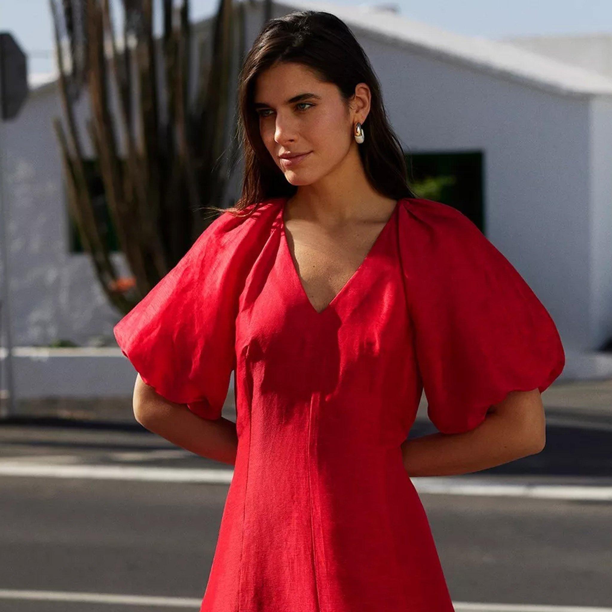A woman in a red dress with puffed sleeves standing outdoors on a street with white buildings and mountains in the background.