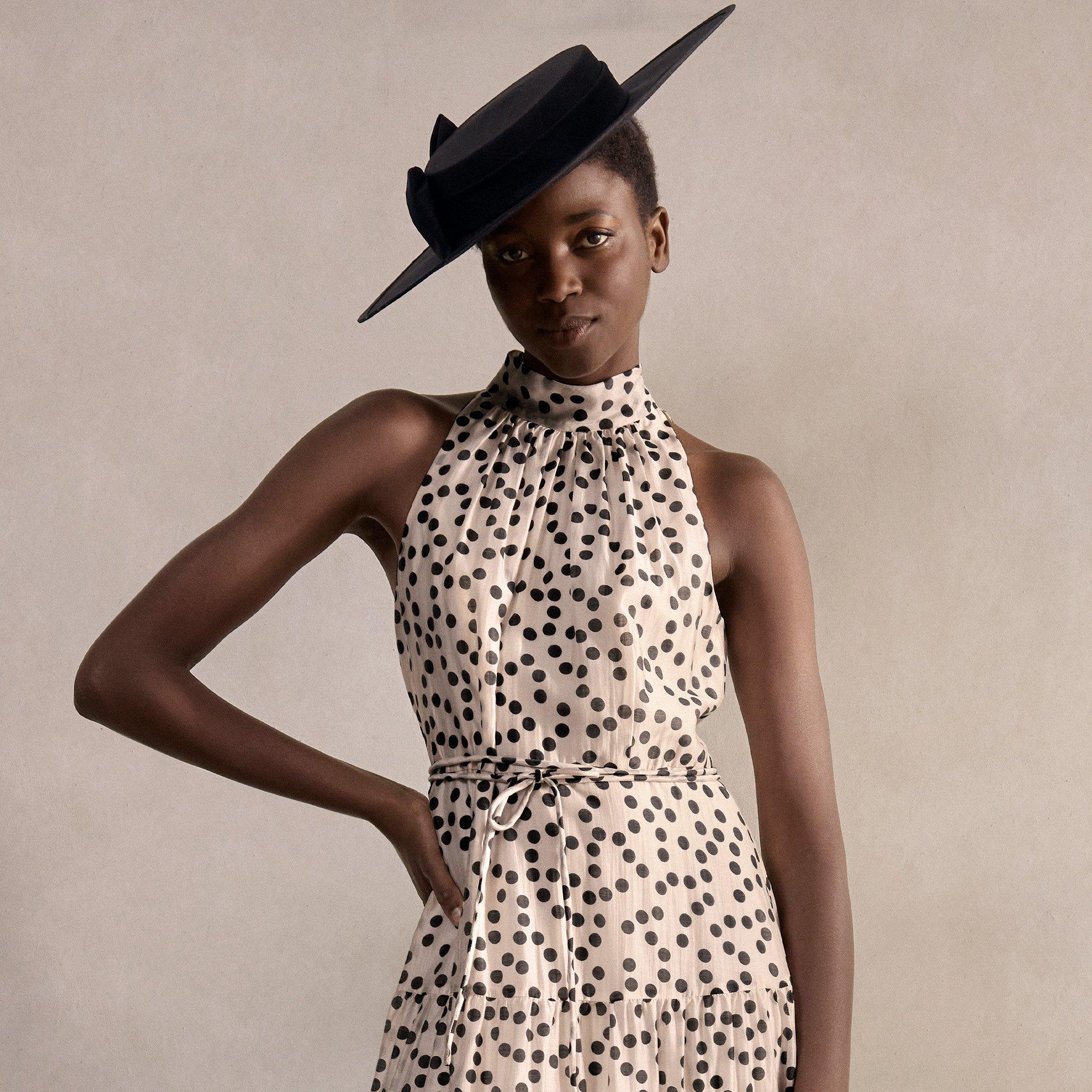 A woman in a polka dot dress with a tied waist, wearing a large black hat, holding a black clutch, standing against a neutral background.