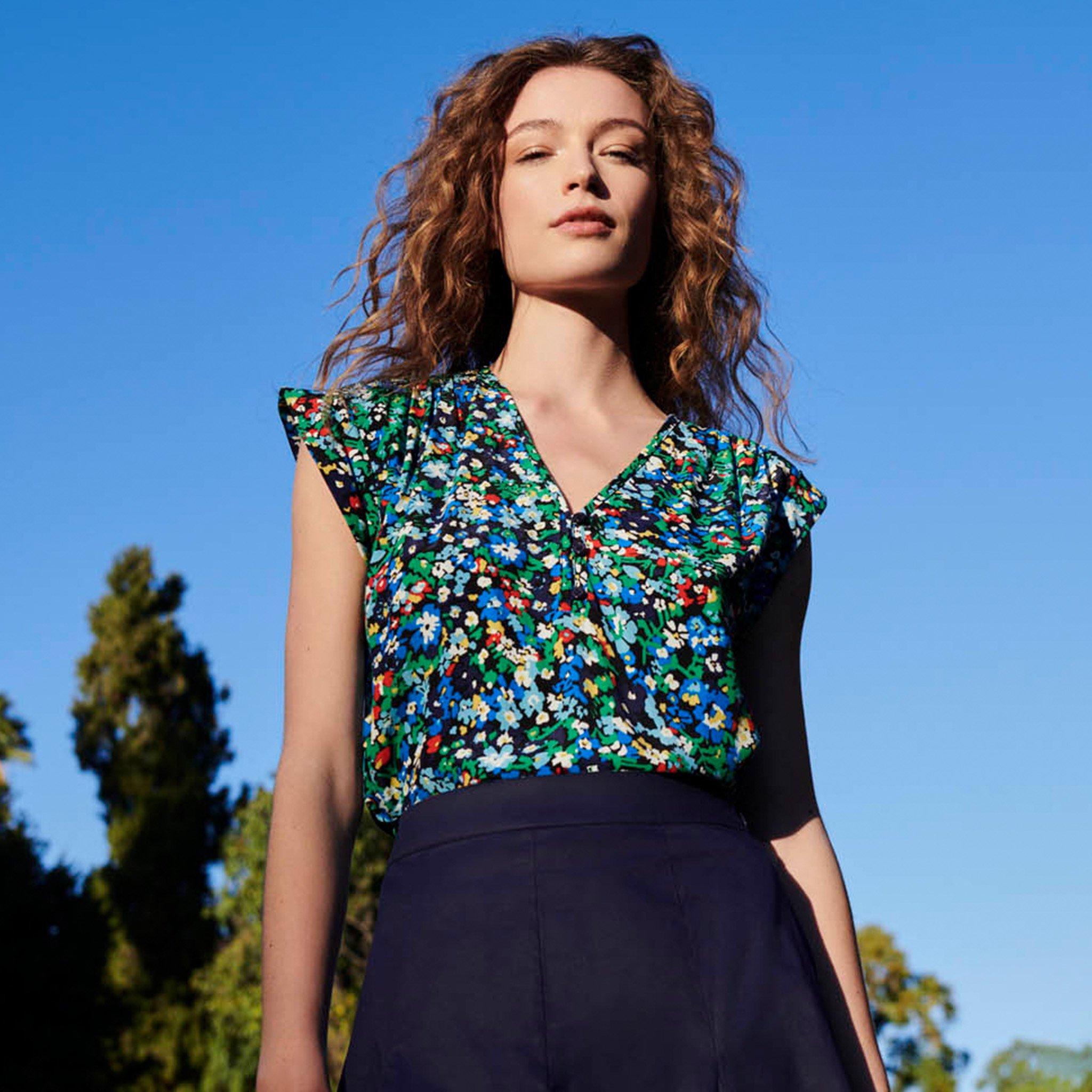 A woman with curly hair wearing a colorful floral top and a dark skirt, standing outdoors against a blue sky.