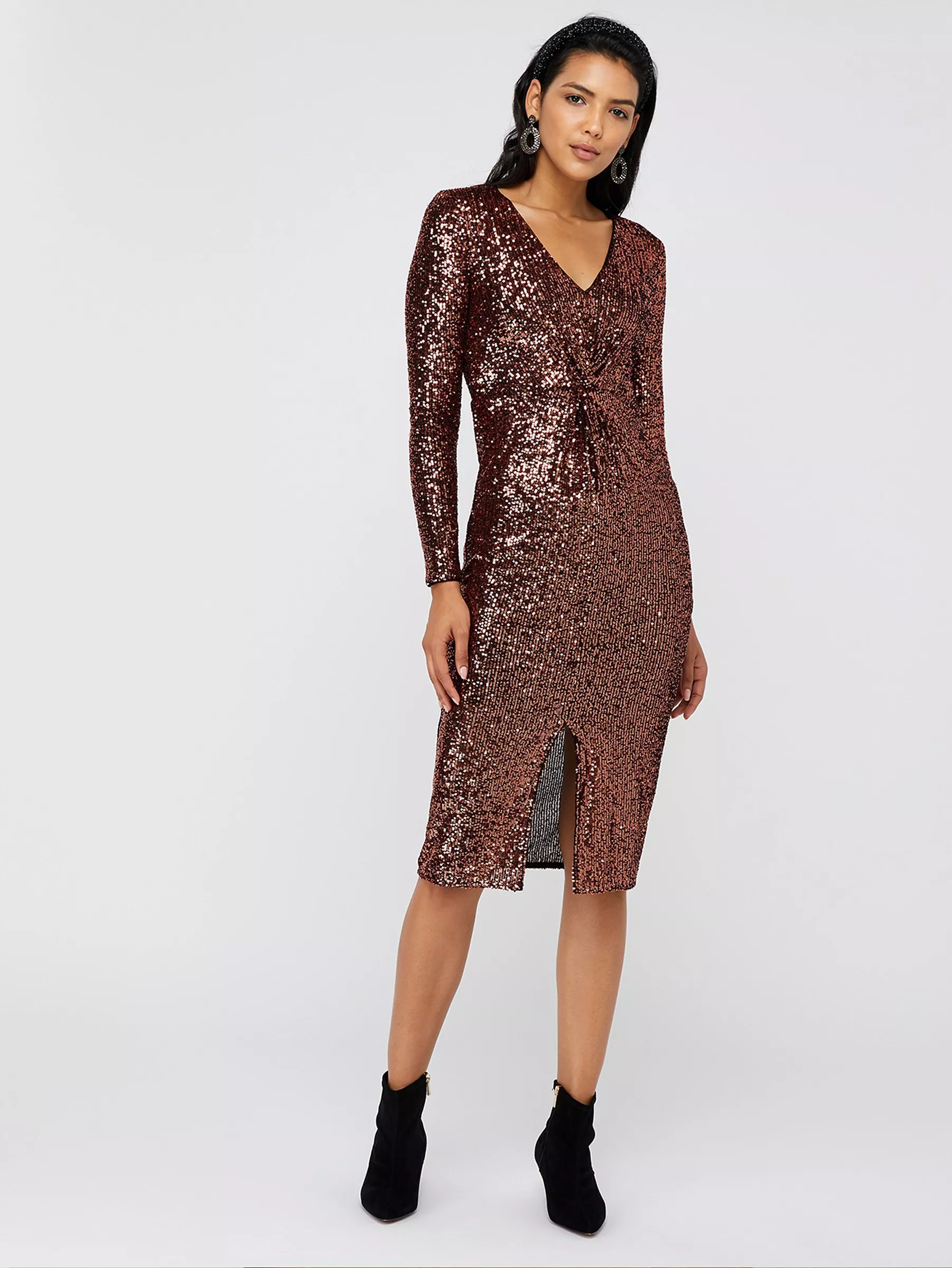 Model wearing a rose gold sequin midi dress with V-neck, long sleeves and front slit, styled with black boots