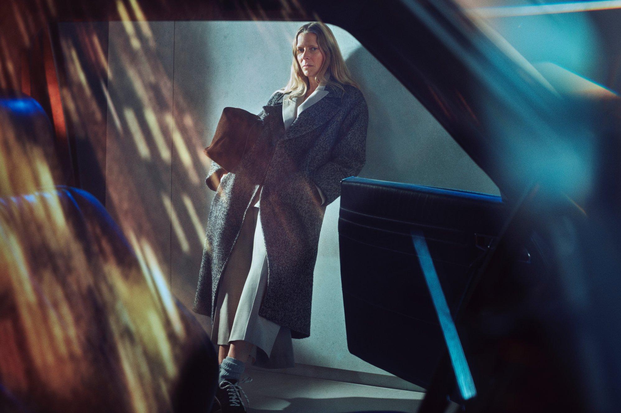 a shot taken through a blurry car door of a women wearing smart trousers and a big trench coat leaning against a wall