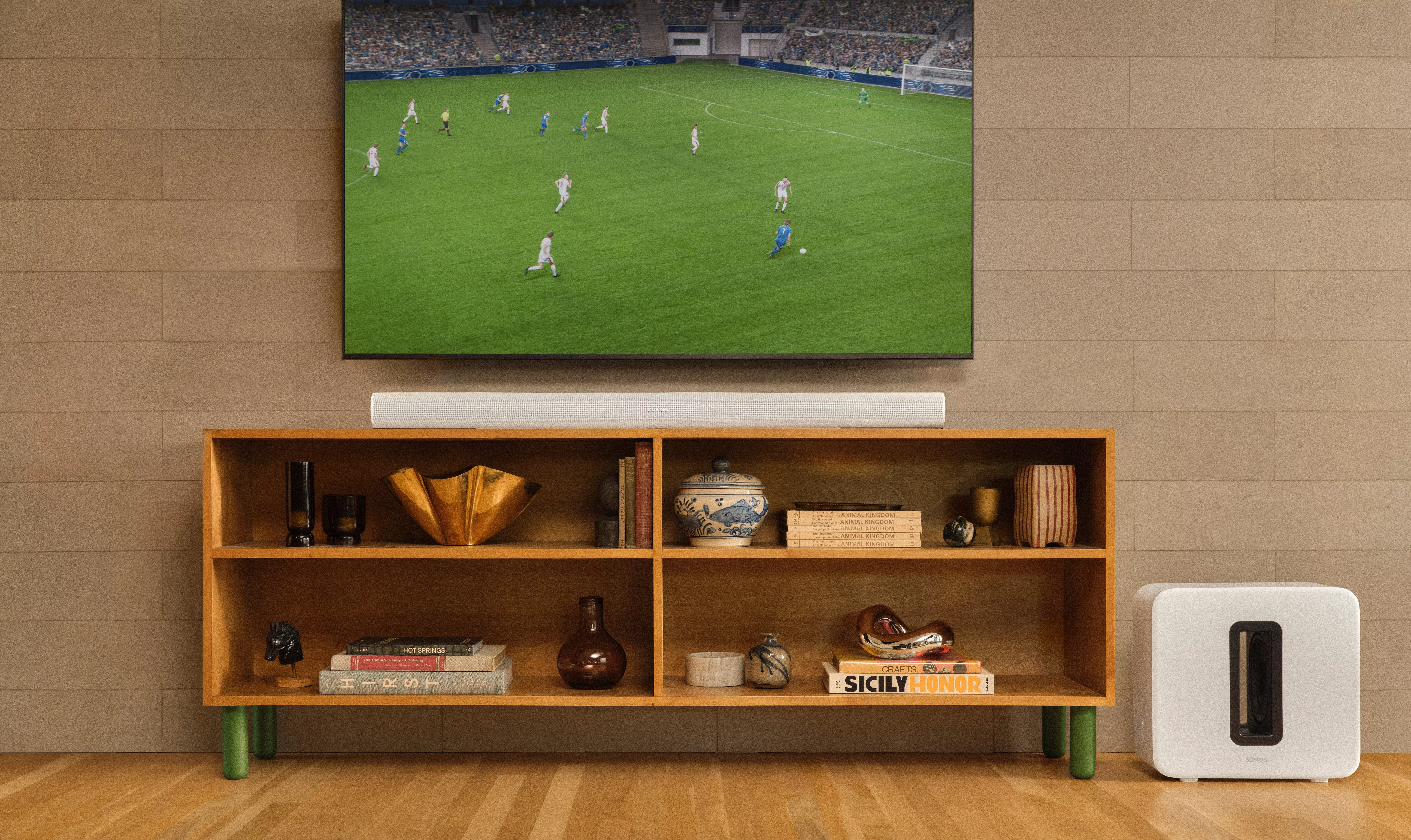 Wall-mounted TV showing a football match above a Sonos soundbar, with Sonos subwoofer beside a wooden media unit.
