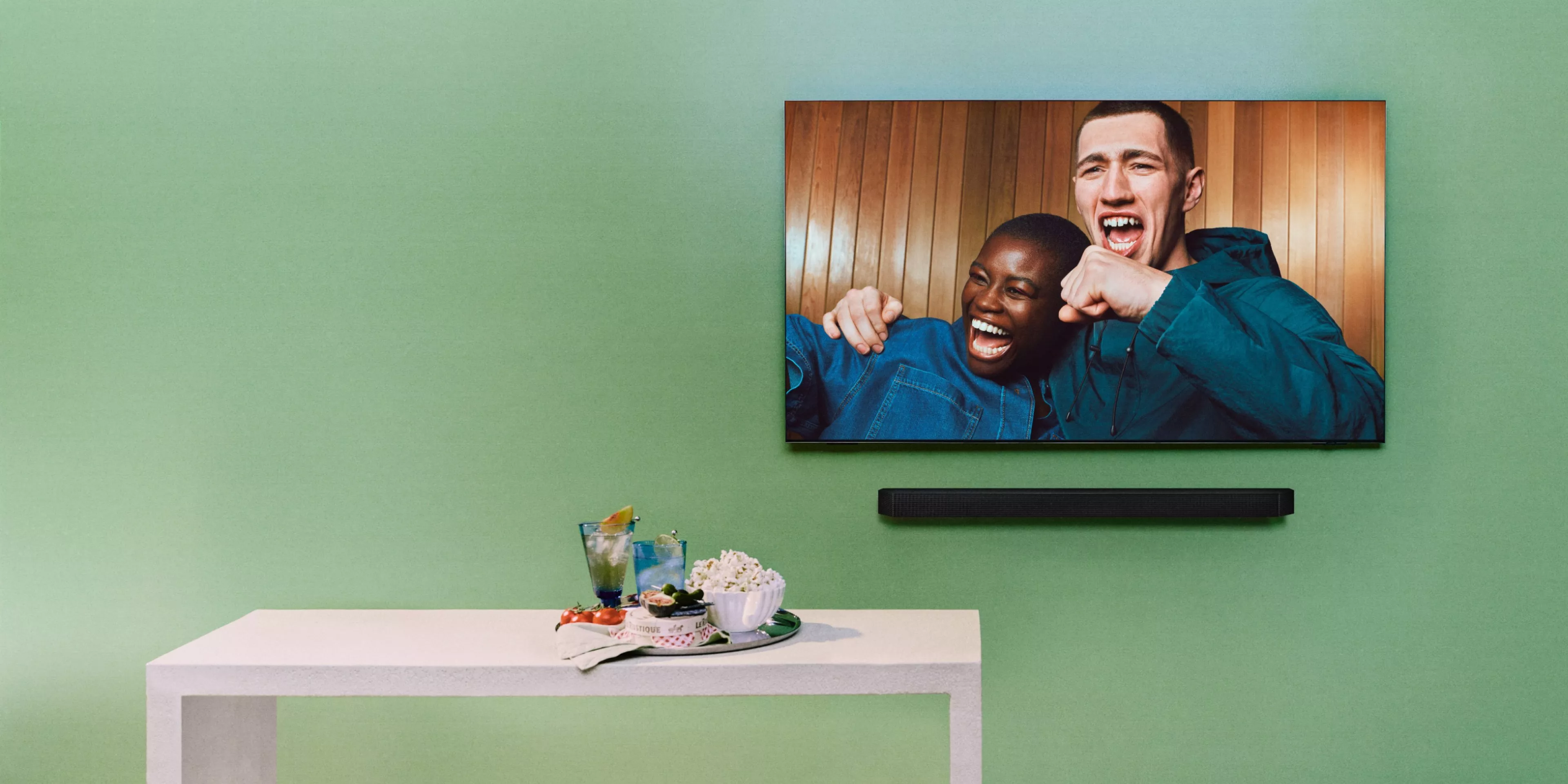 tv on a green wall with 2 people chearing on the TV with a tray of drinks and snacks below