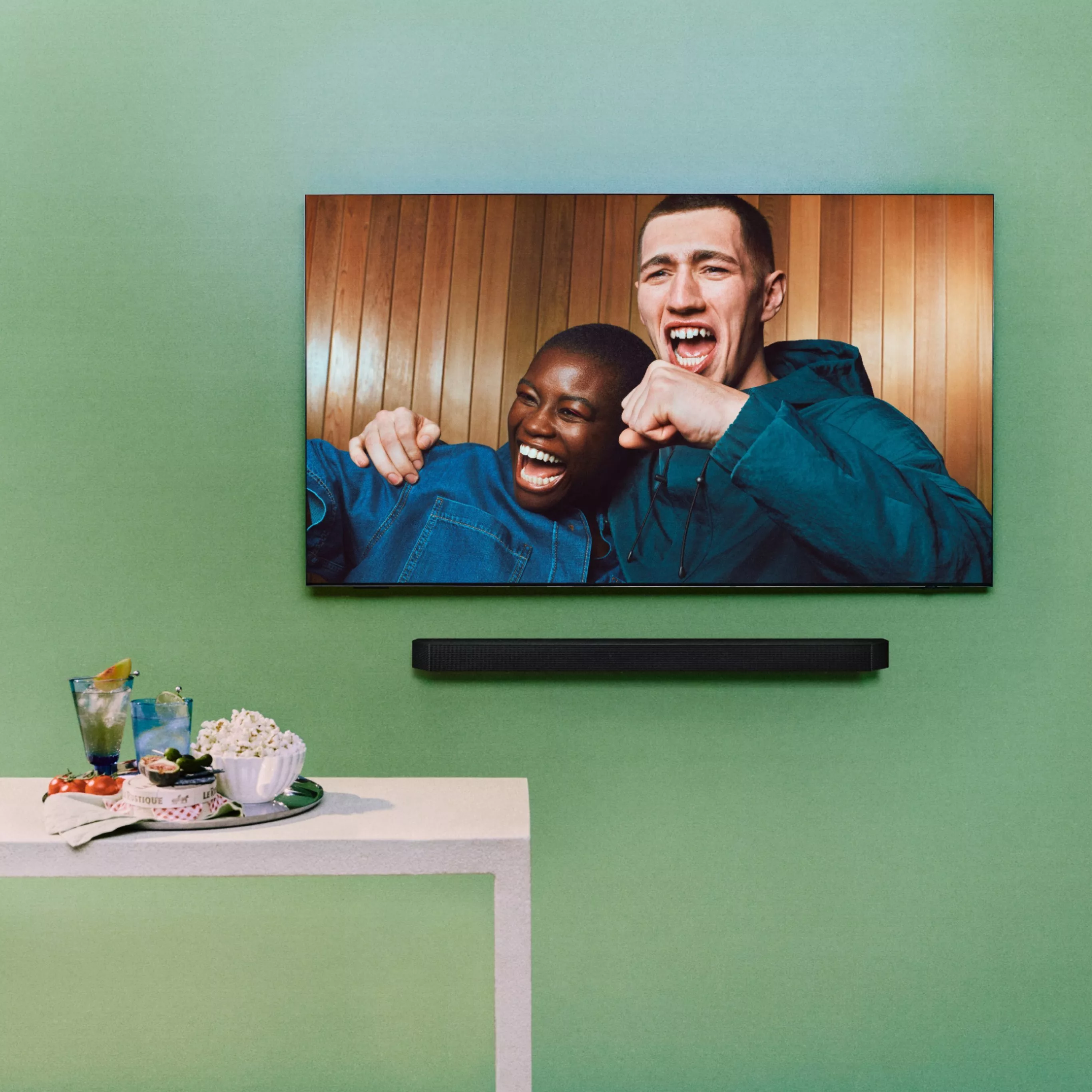 Wall-mounted TV showing two friends cheering, with a soundbar and snacks on a table beneath the screen