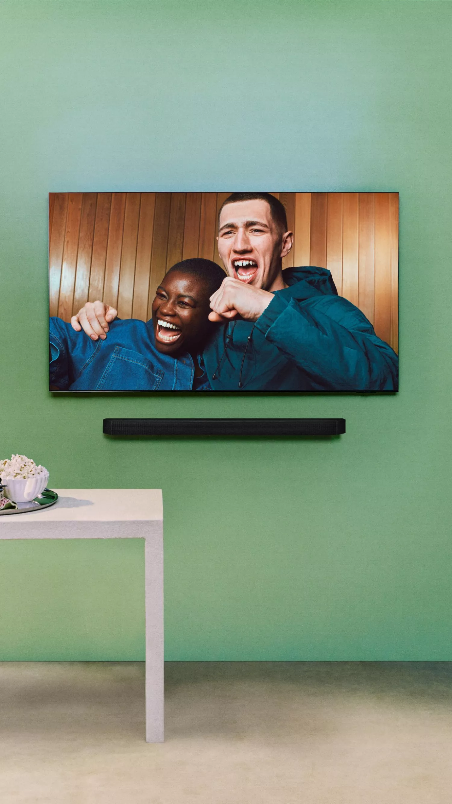 tv on a green wall with 2 people chearing on the TV with a tray of drinks and snacks below