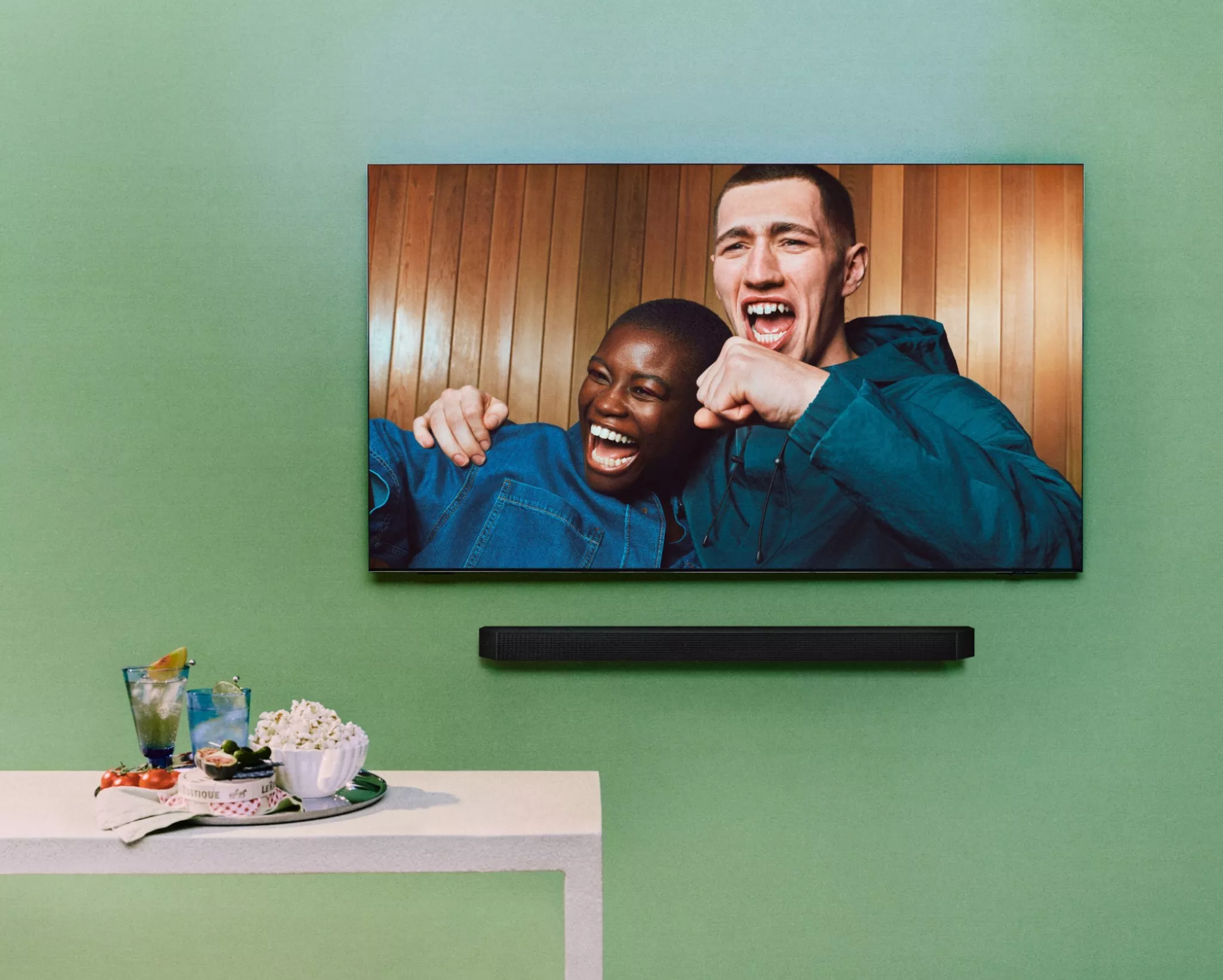 tv mounted on a green wall with a man and women chearing on the screen with a try of drinks and snacks below