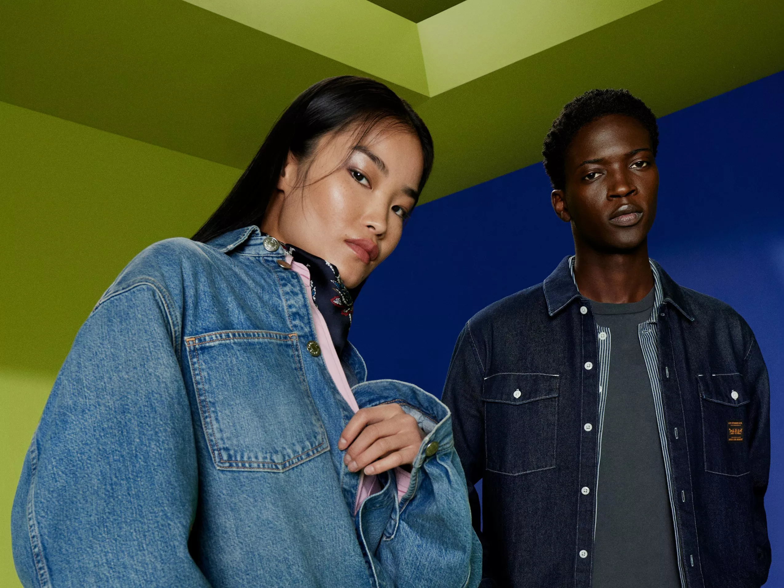 Two fashion models in denim jackets posing indoors against a bold green and blue geometric background