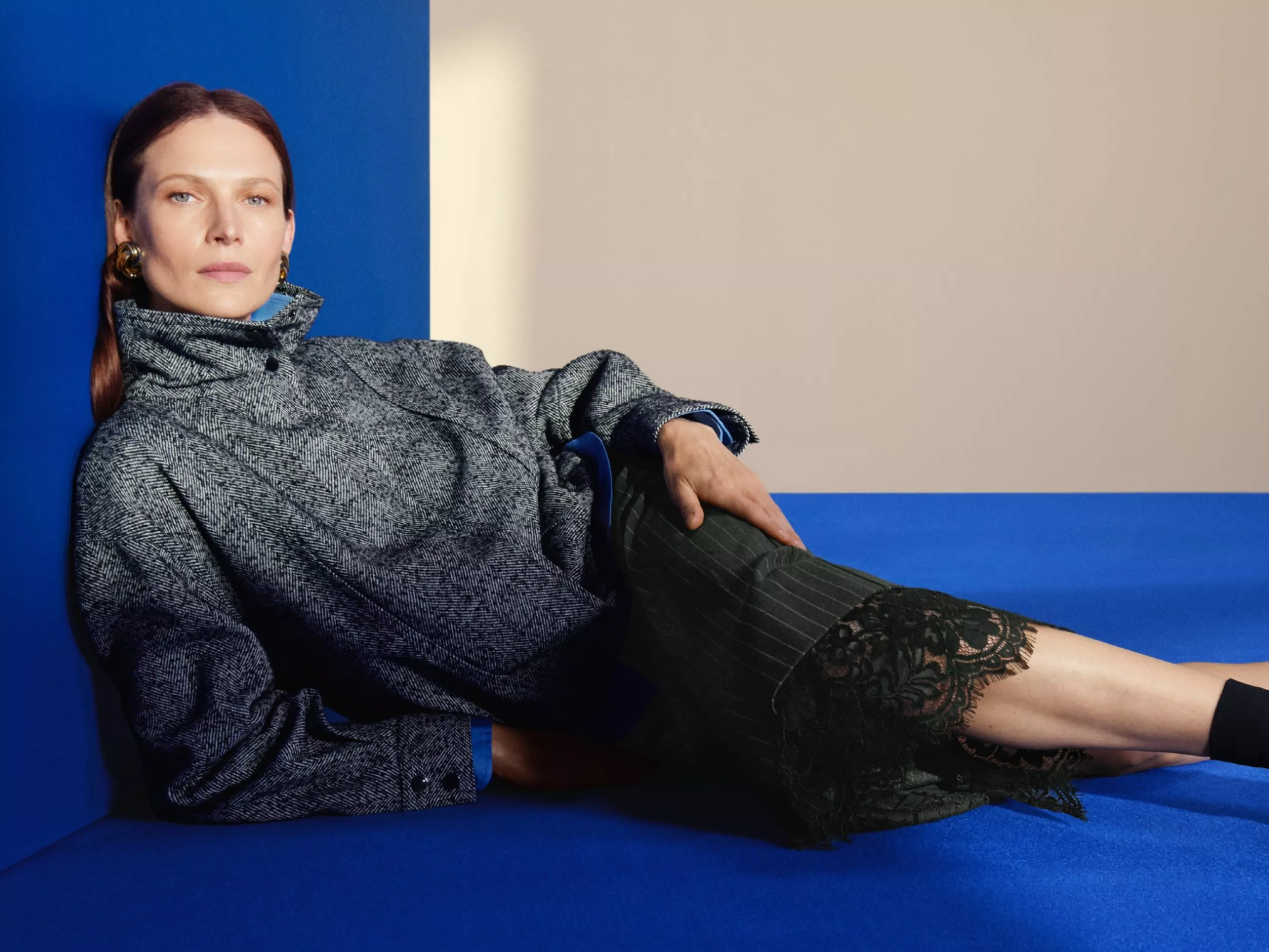 Fashion portrait of a woman reclining on a blue sofa, wearing a grey high-neck coat and lace skirt.