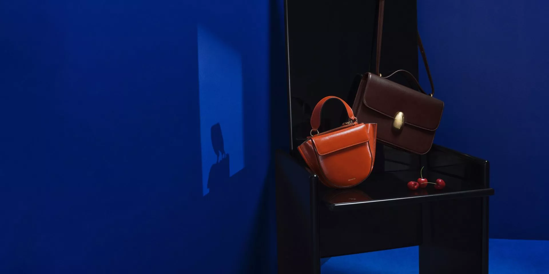 Luxury leather handbags on a black chair with cherries, set against a deep blue studio background