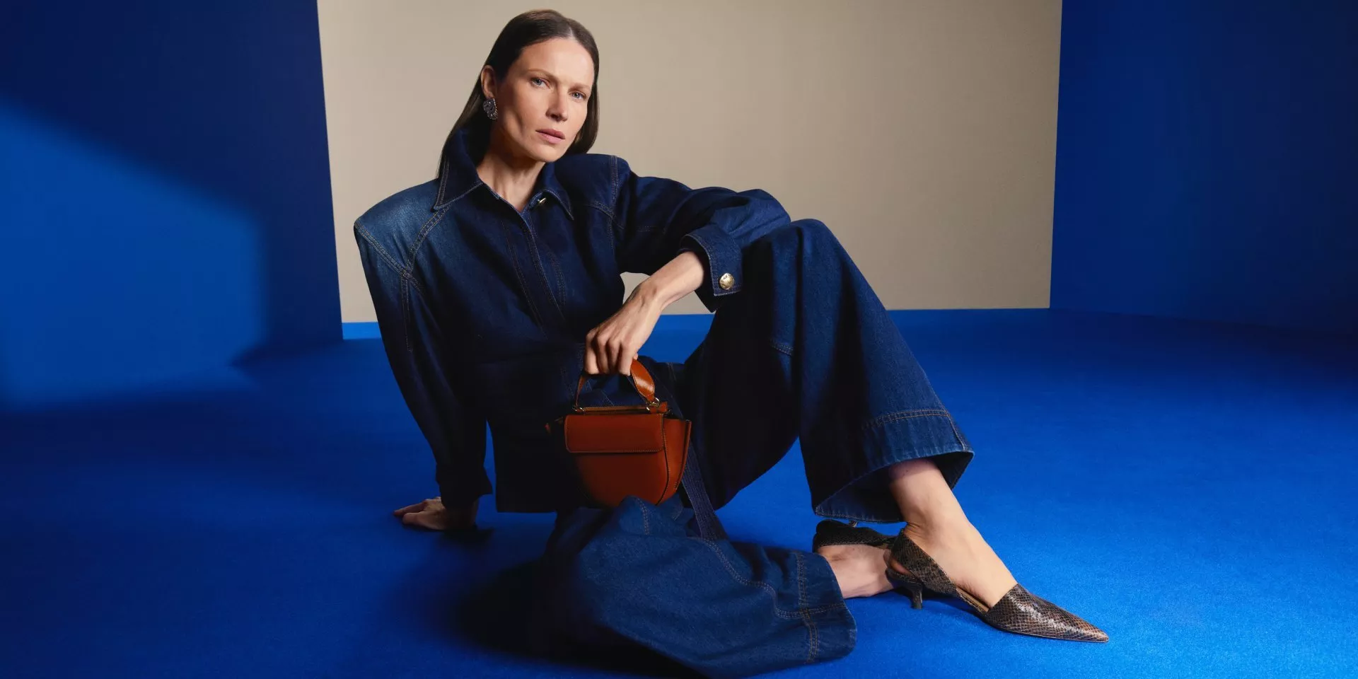 Fashion model in dark denim jumpsuit holding brown handbag, seated on blue studio floor, minimalist backdrop