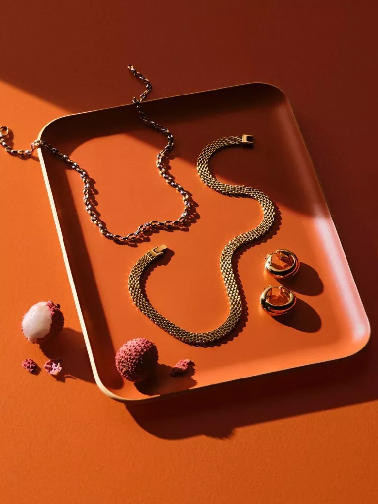Gold chain necklace and hoop earrings on an orange tray, luxury jewellery flat lay with warm light and shadows