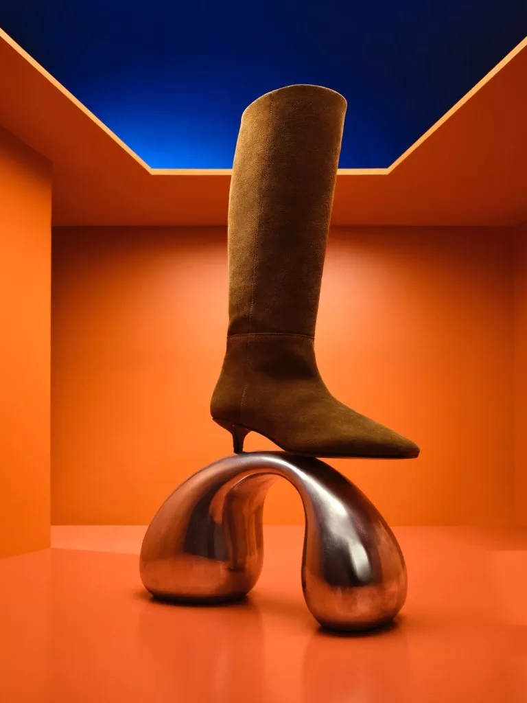 Brown suede knee-high heeled boot on a polished metal sculpture in a bright orange room with blue ceiling
