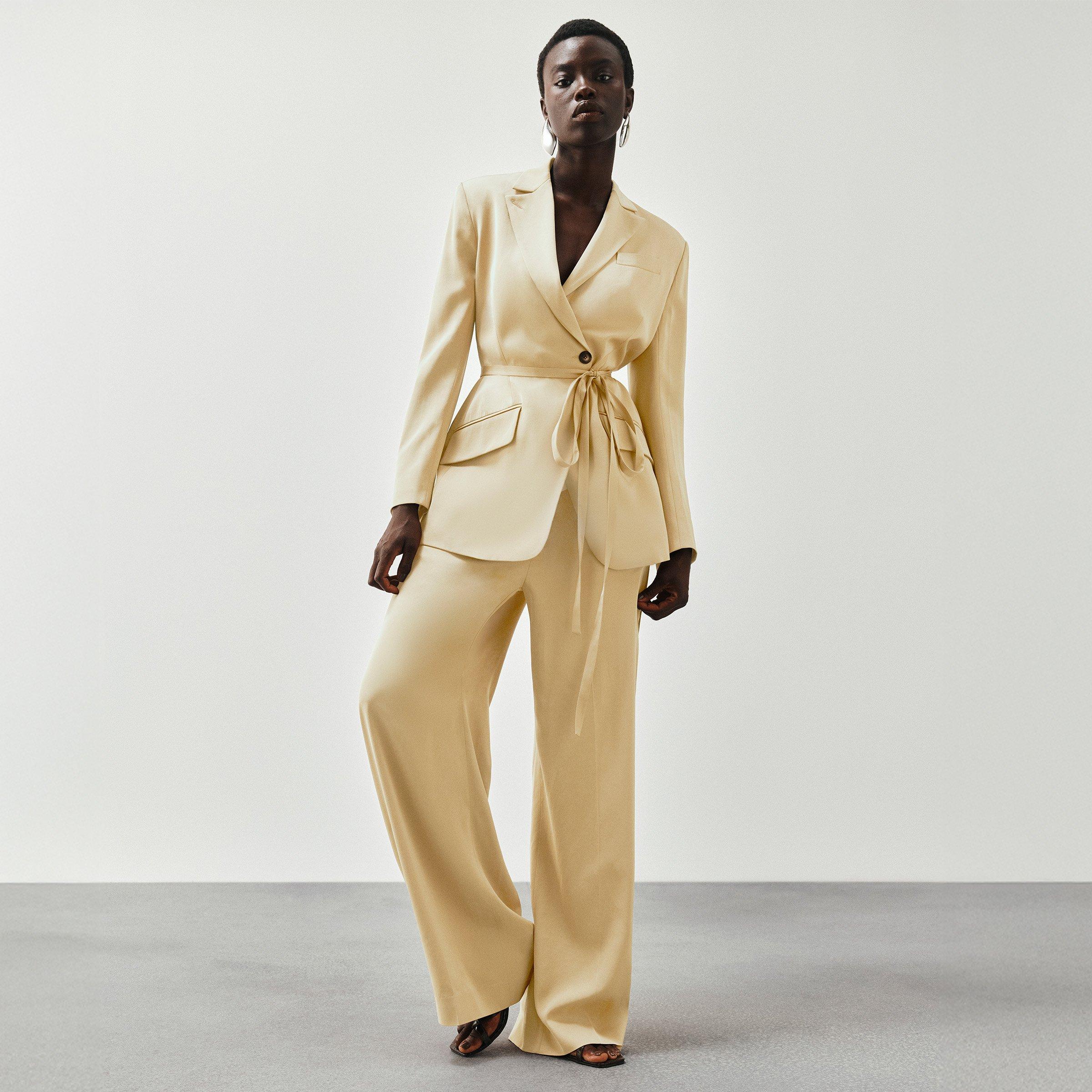 Woman in a beige pantsuit with a belted blazer, standing confidently against a plain background.