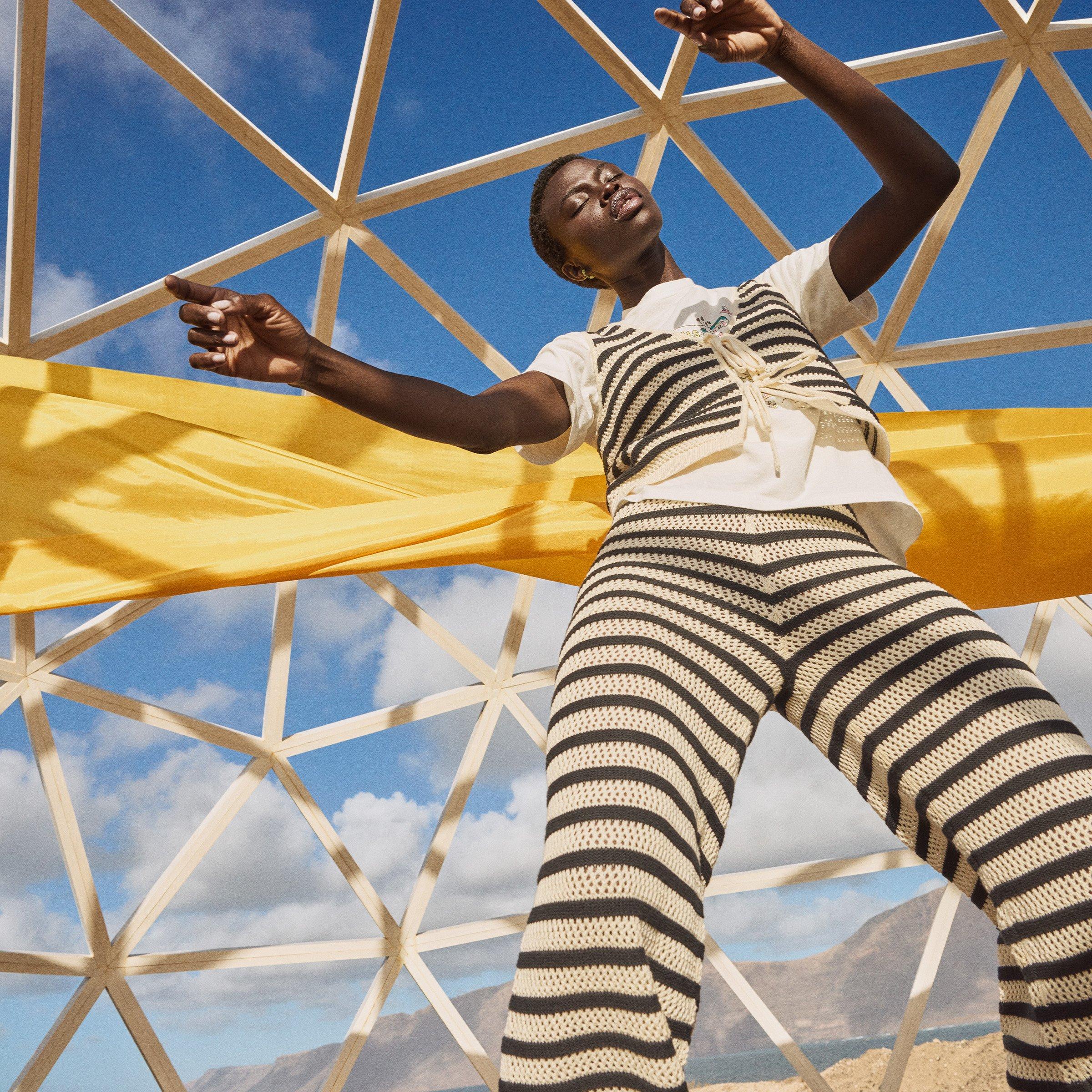 Model poses under a geodesic dome wearing a striped crochet outfit with a yellow fabric backdrop and blue sky.