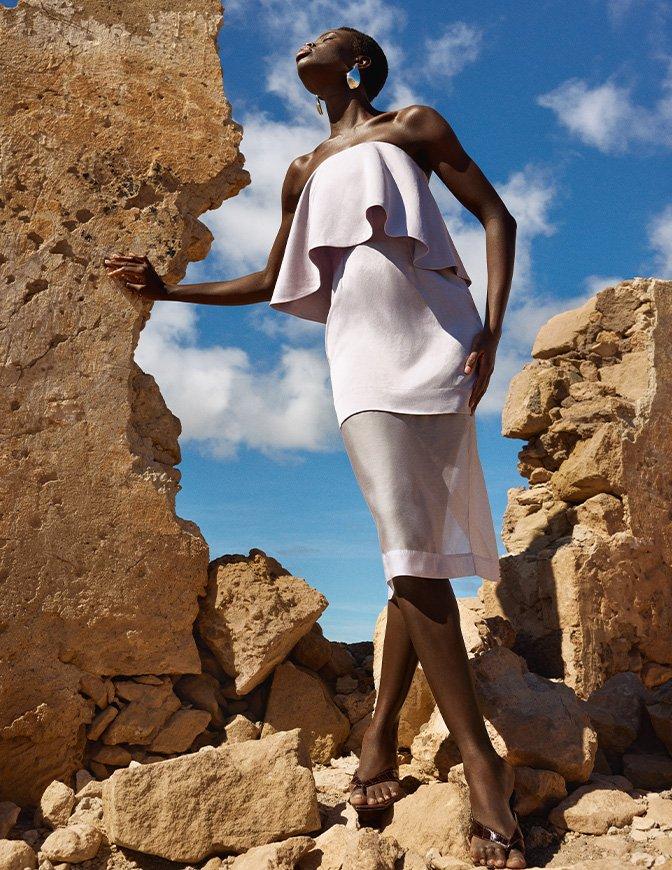 Woman posing confidently among rocky ruins, wearing a strapless white ruffled dress and heeled sandals, with a bright blue sky in the background.