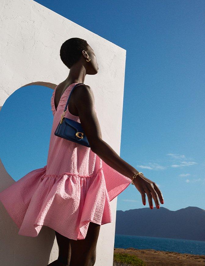 Woman in a textured pink dress and gold accessories poses outdoors with a blue shoulder bag, set against a scenic ocean and mountain backdrop.