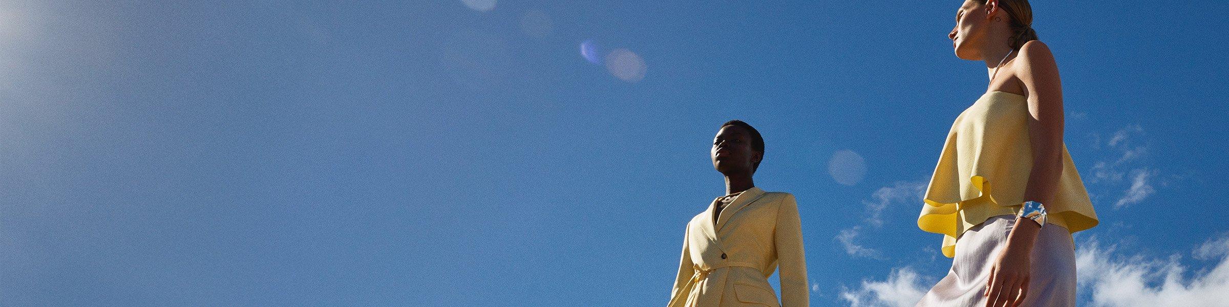 Two women against blue sky, one woman in yellow jacket, and another in yellow flowing top.