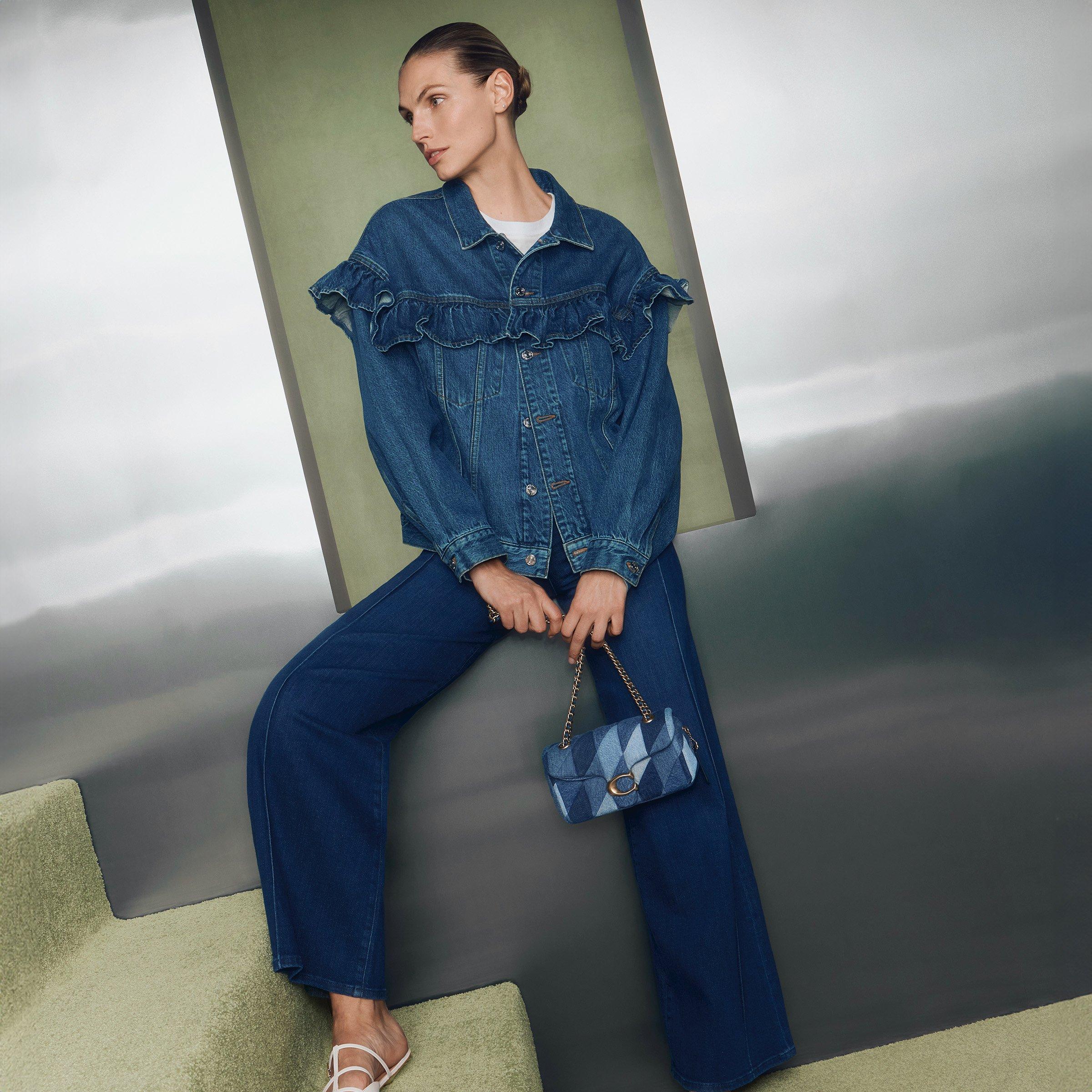Woman standing in stairs wearing in denim jacket and wide legged trousers, carrying denim handbag.