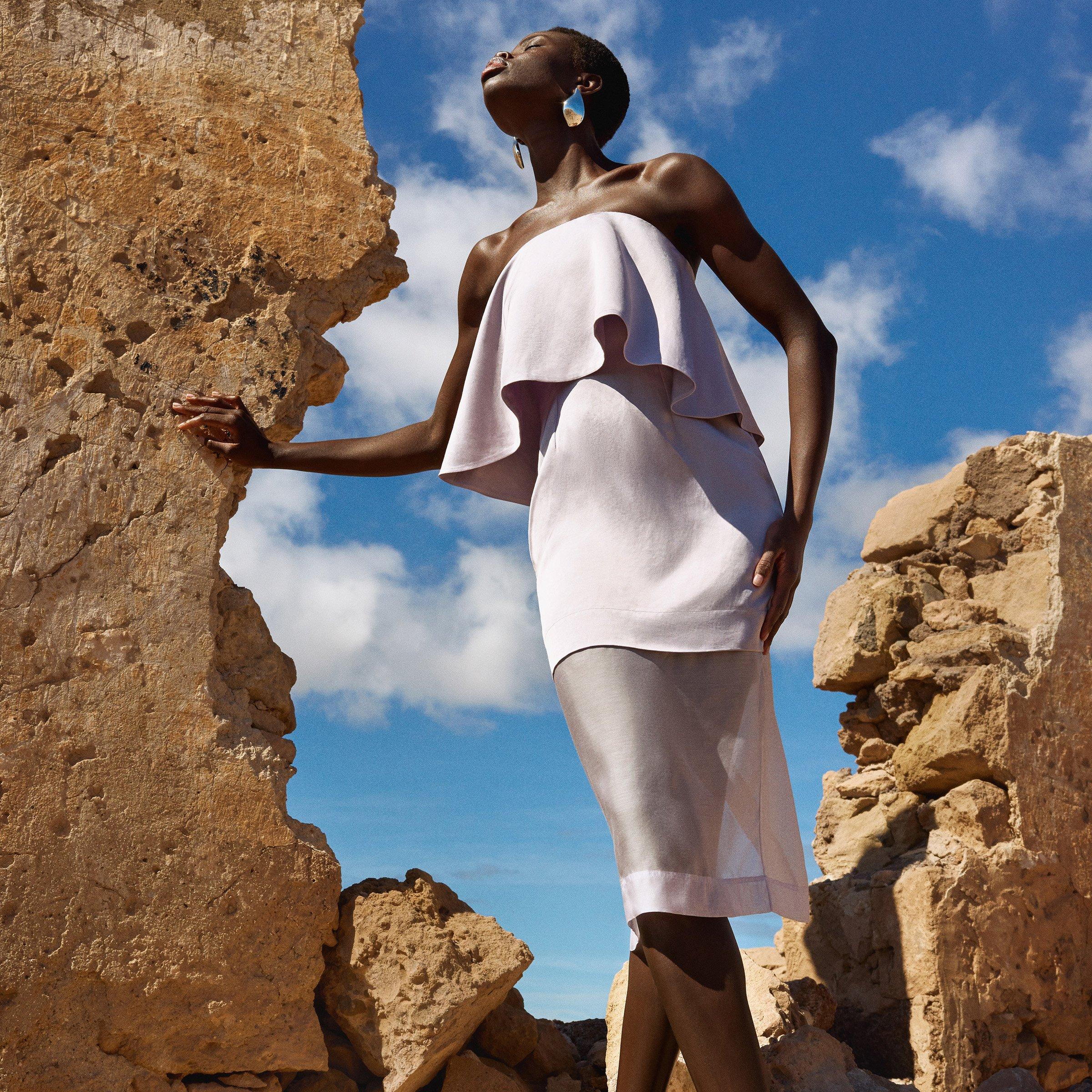 Woman wearing white strapless dress, looking up to the sky against rocky backdrop.
