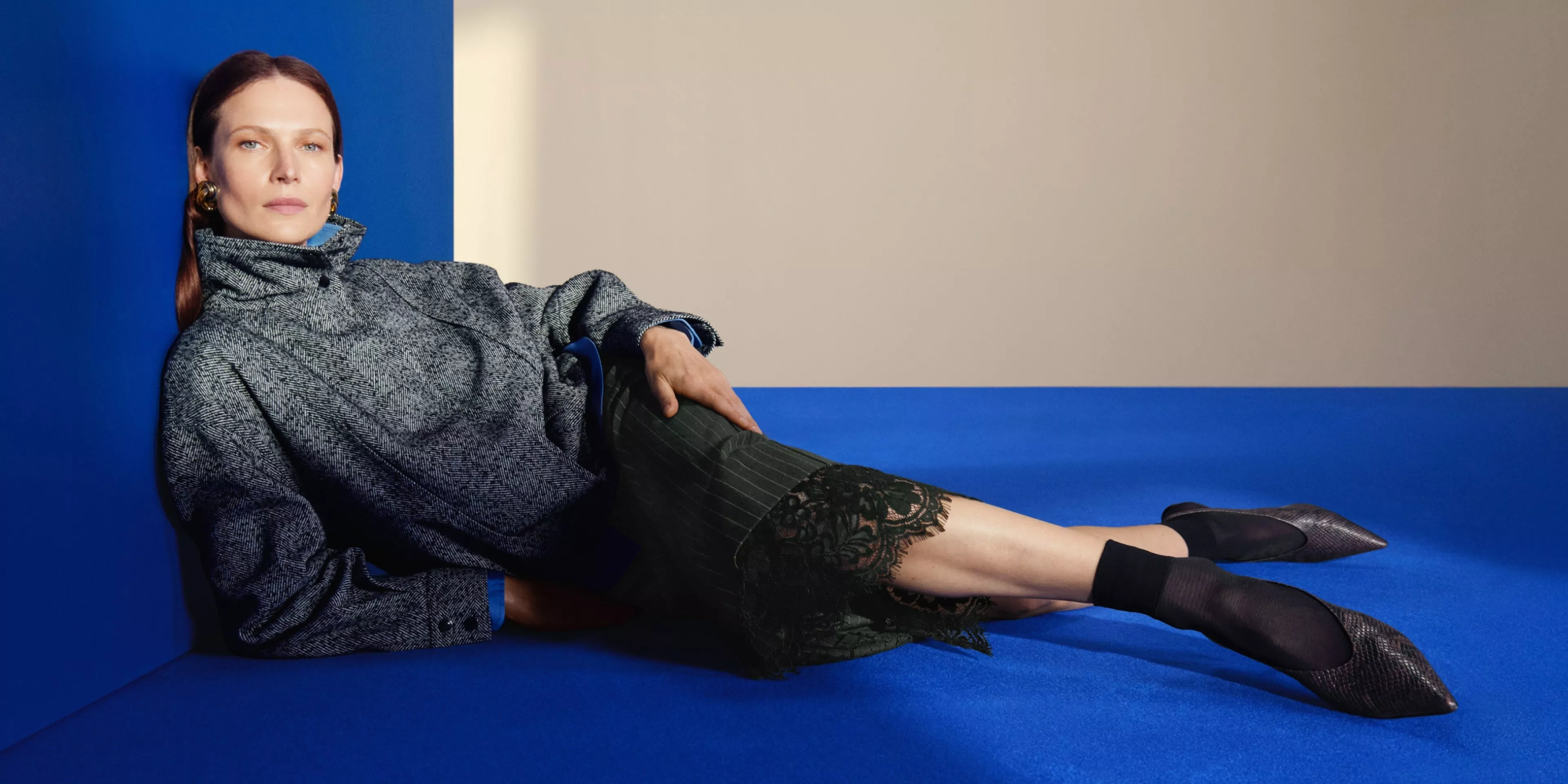 Fashion model reclining on a blue sofa, wearing a grey high-neck jacket and lace-trim skirt in studio setting
