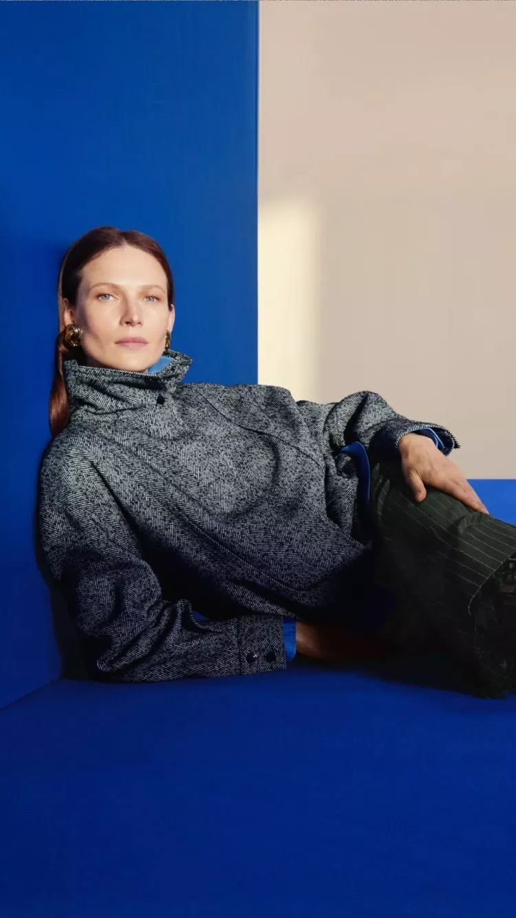 Fashion model reclining on cobalt blue floor, wearing grey high-neck jacket, lace skirt and heels