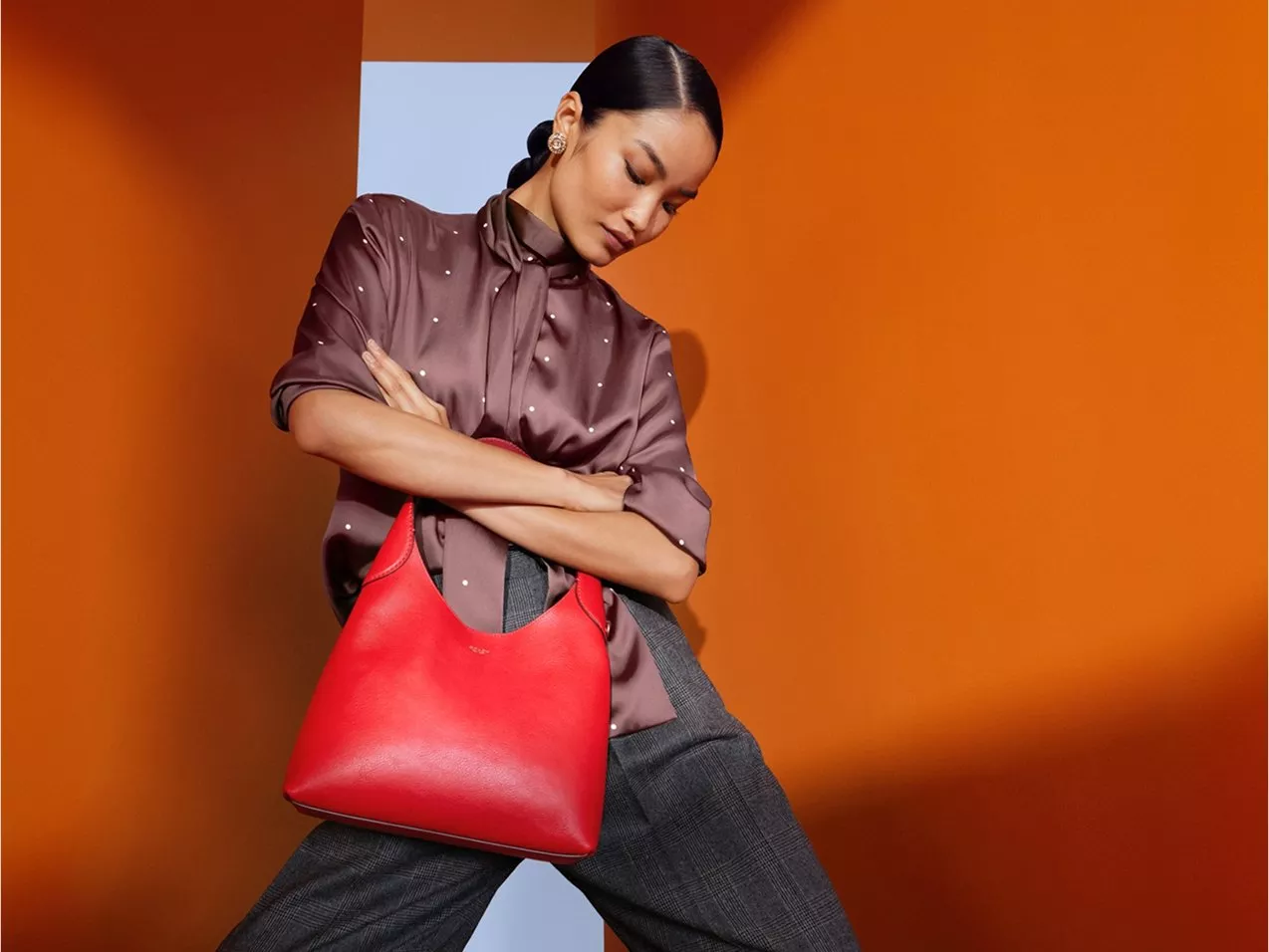 Fashion model holding red leather hobo handbag, wearing satin blouse and trousers against orange backdrop