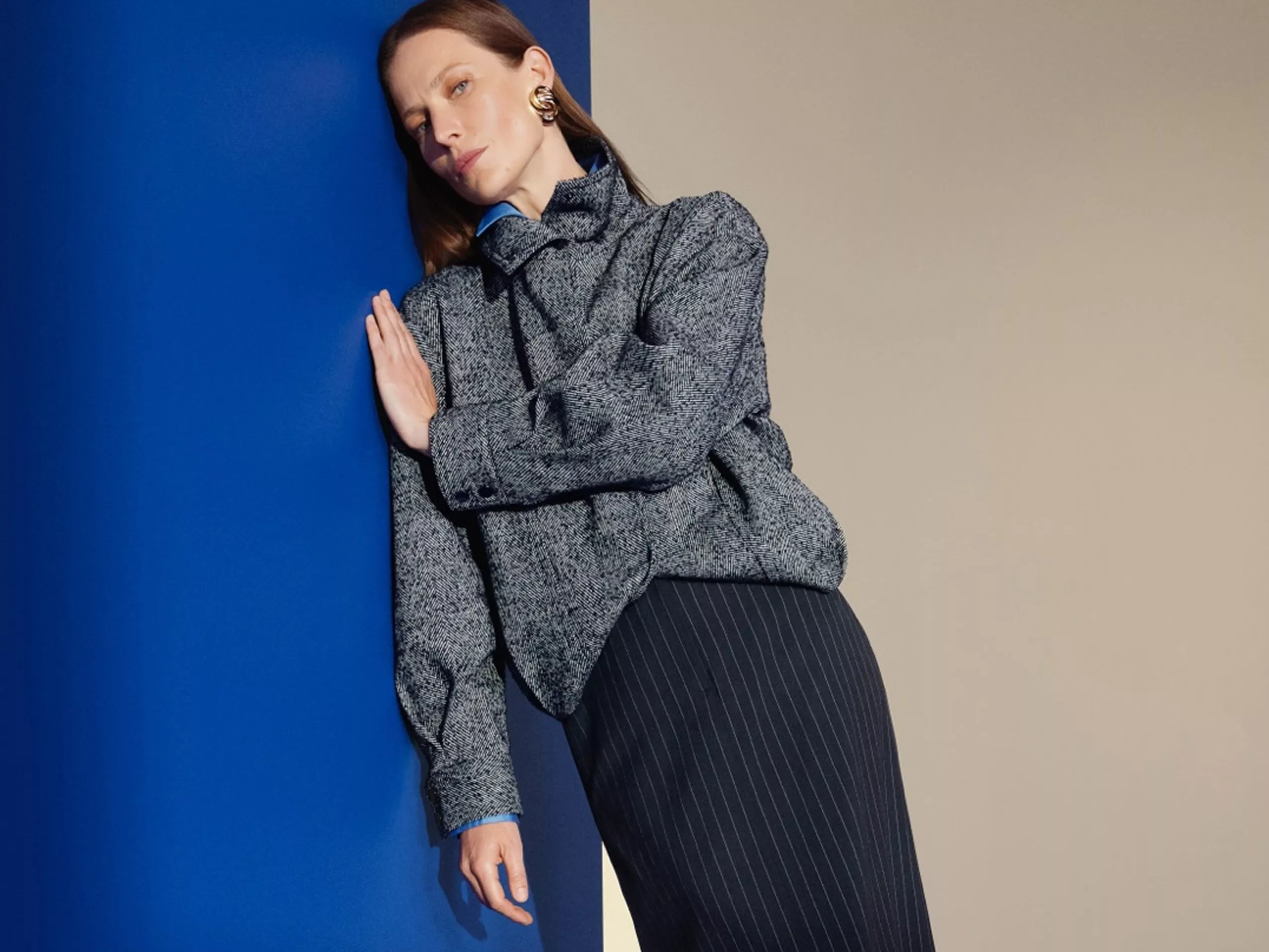 Fashion model in grey tweed jacket and pinstripe skirt leaning against blue and beige wall