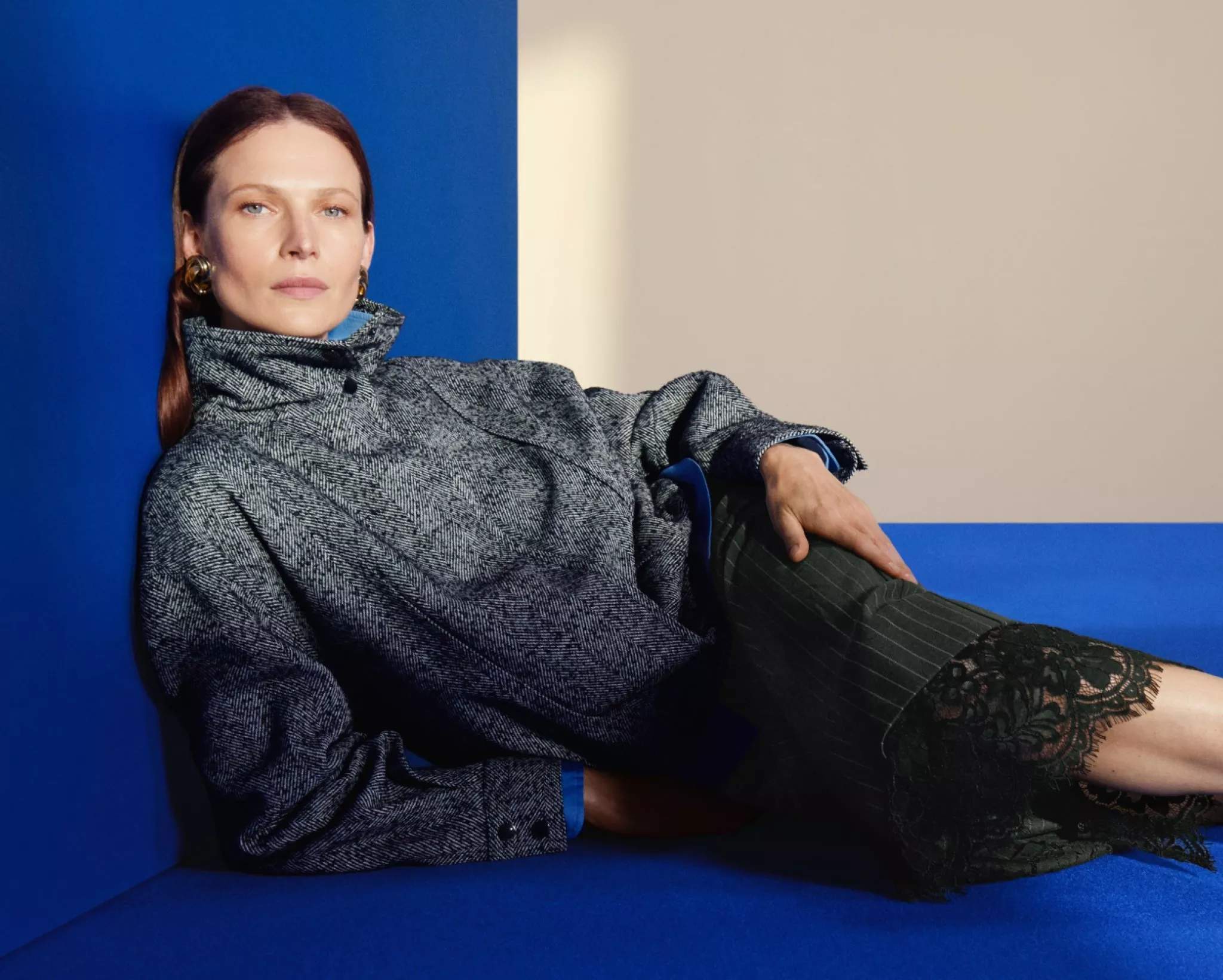 Fashion portrait of woman reclining on blue flooring, wearing grey coat and black lace trim skirt
