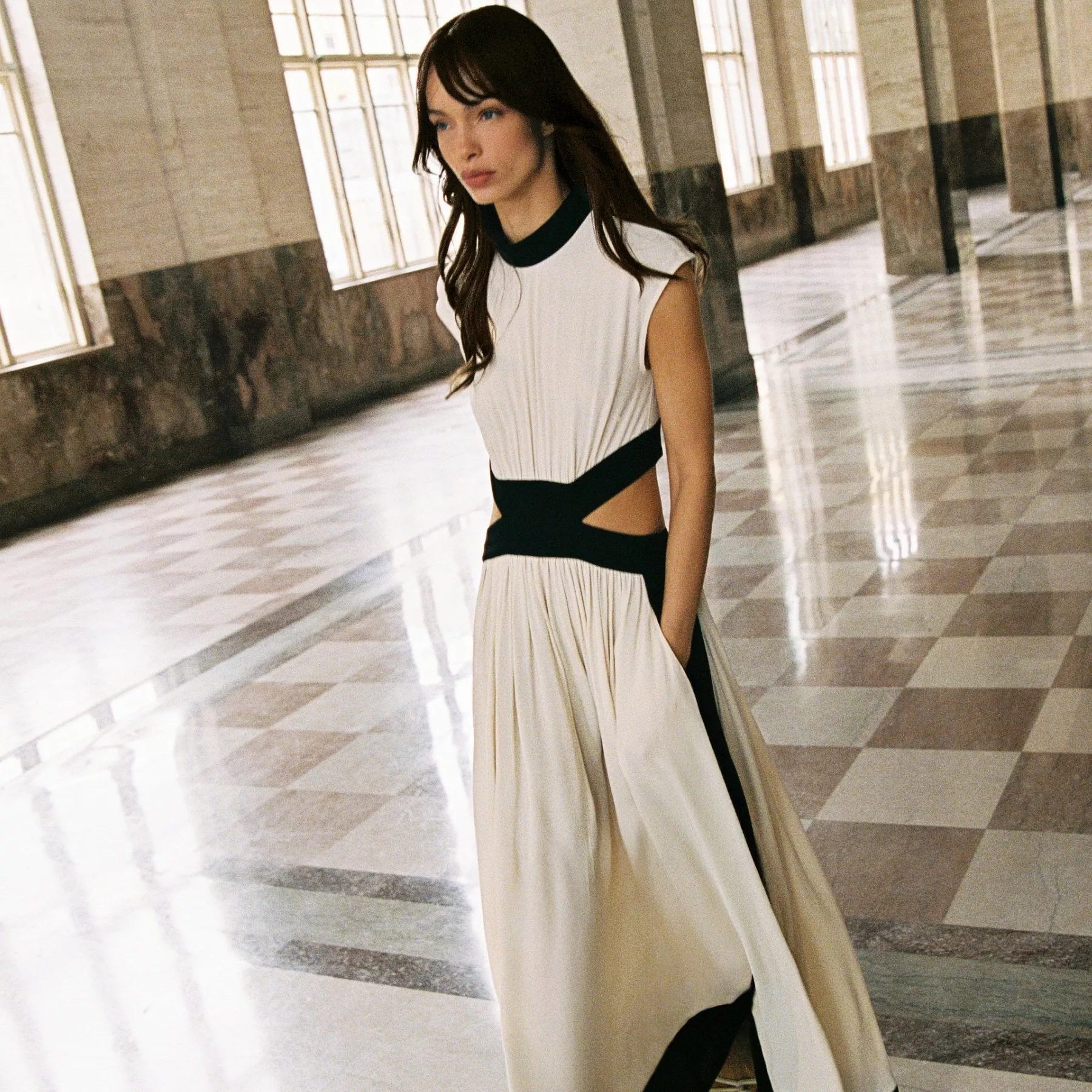 Elegant woman in a flowing white dress with black accents walks through a spacious, sunlit marble hall.