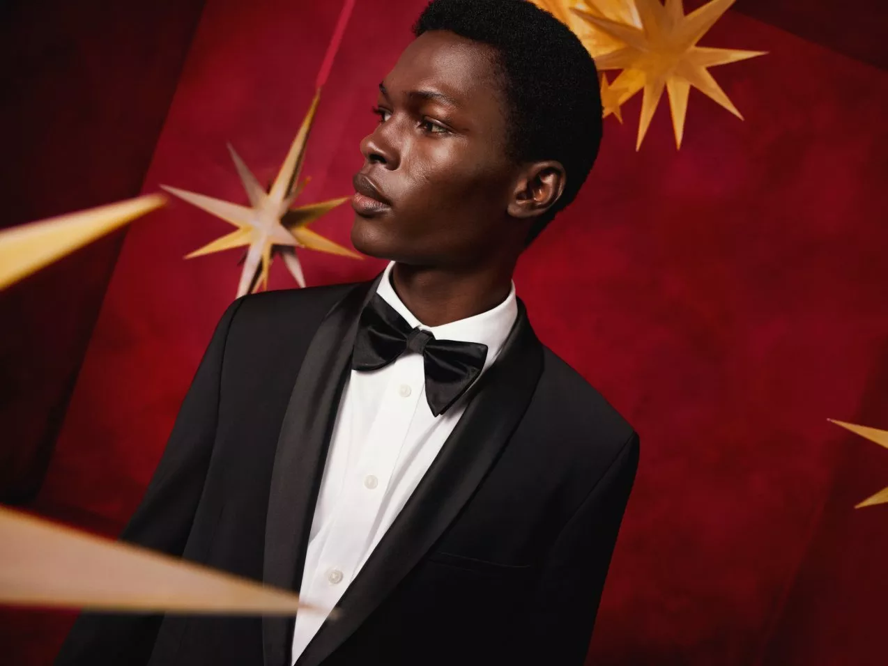 Young man in black tuxedo and bow tie against red backdrop with gold stars, elegant formal fashion portrait