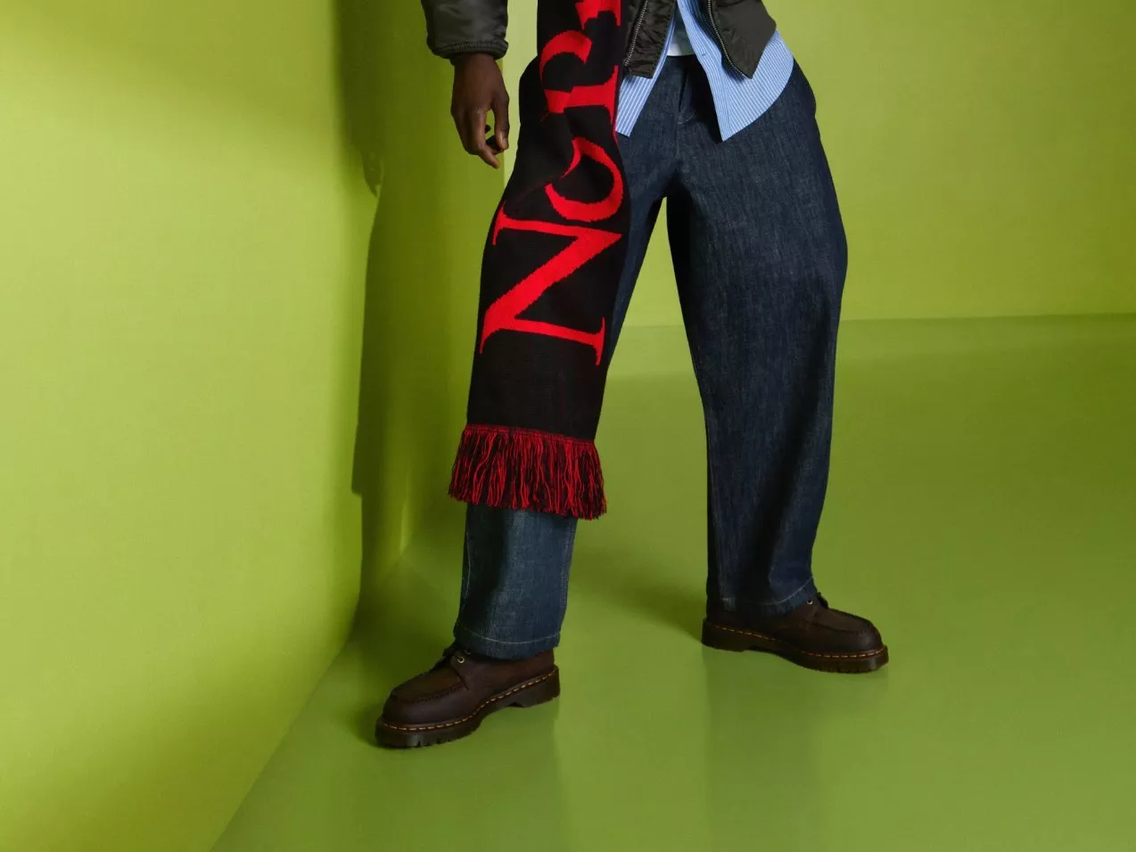 Fashion model wearing dark jeans, leather jacket and bold red-letter scarf, standing in green studio backdrop
