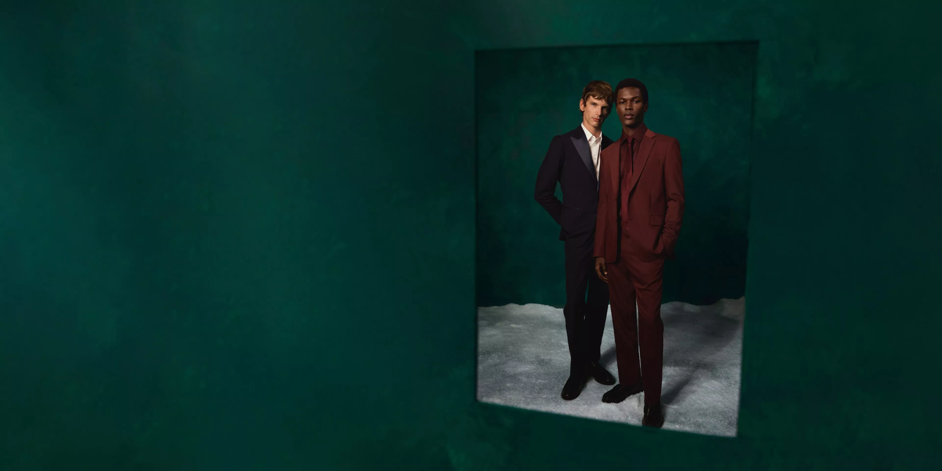 Two male models in tailored suits posing in a studio with dark green backdrop, showcasing formal menswear fashion