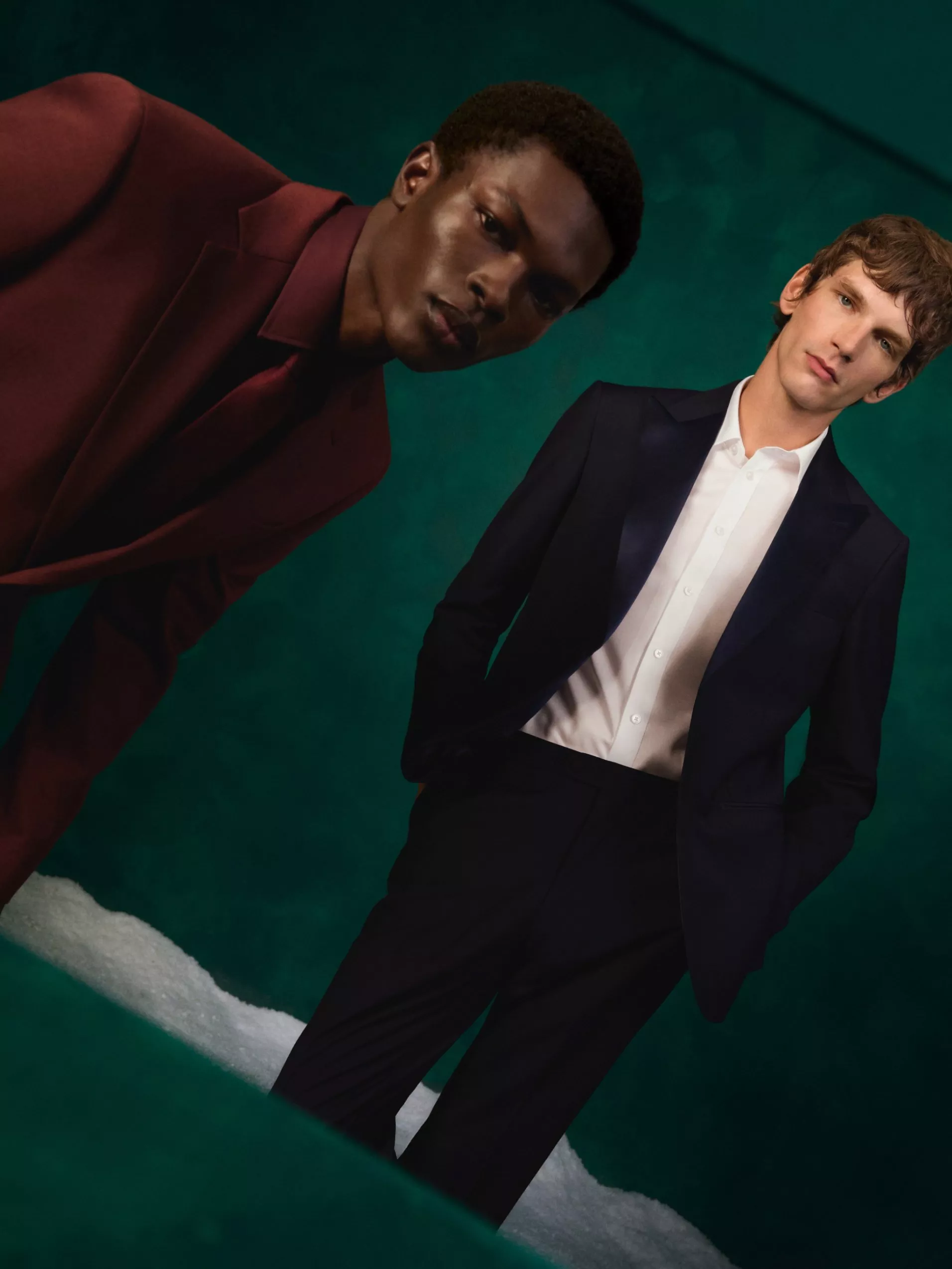 Two male models in tailored burgundy and navy suits posing against a teal studio background, fashion editorial portrait