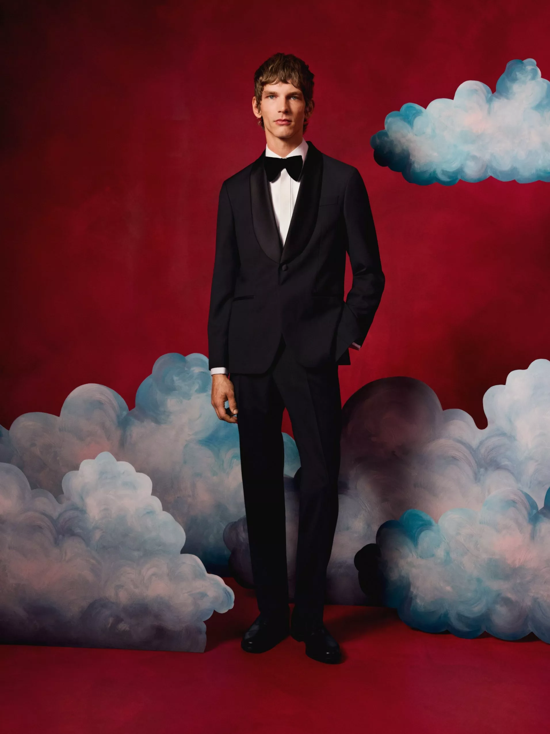 Man in black tuxedo and bow tie on red studio backdrop with painted clouds, formal menswear fashion shoot