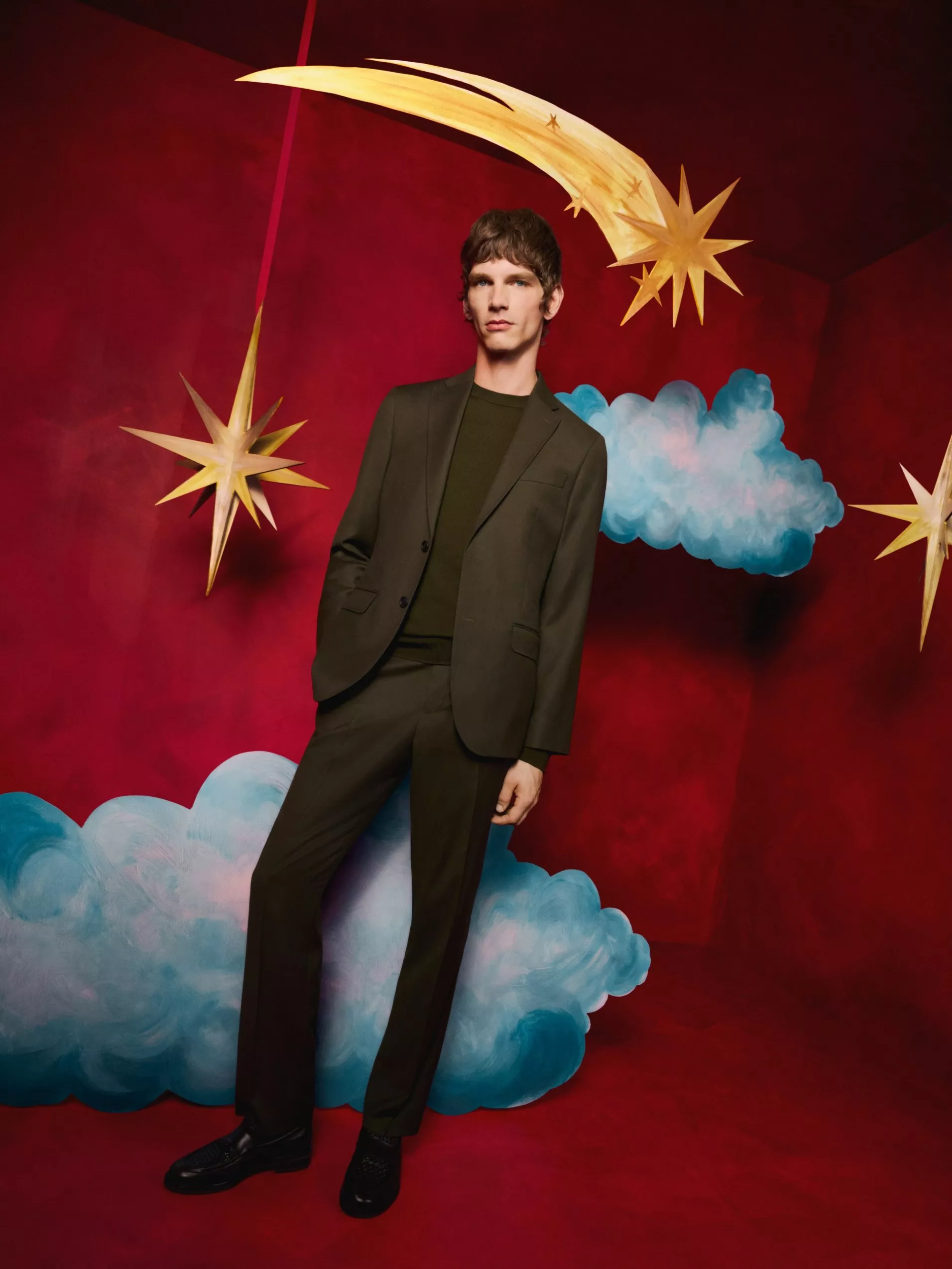 Fashion portrait of a man in an olive suit among clouds and gold stars on a red studio backdrop