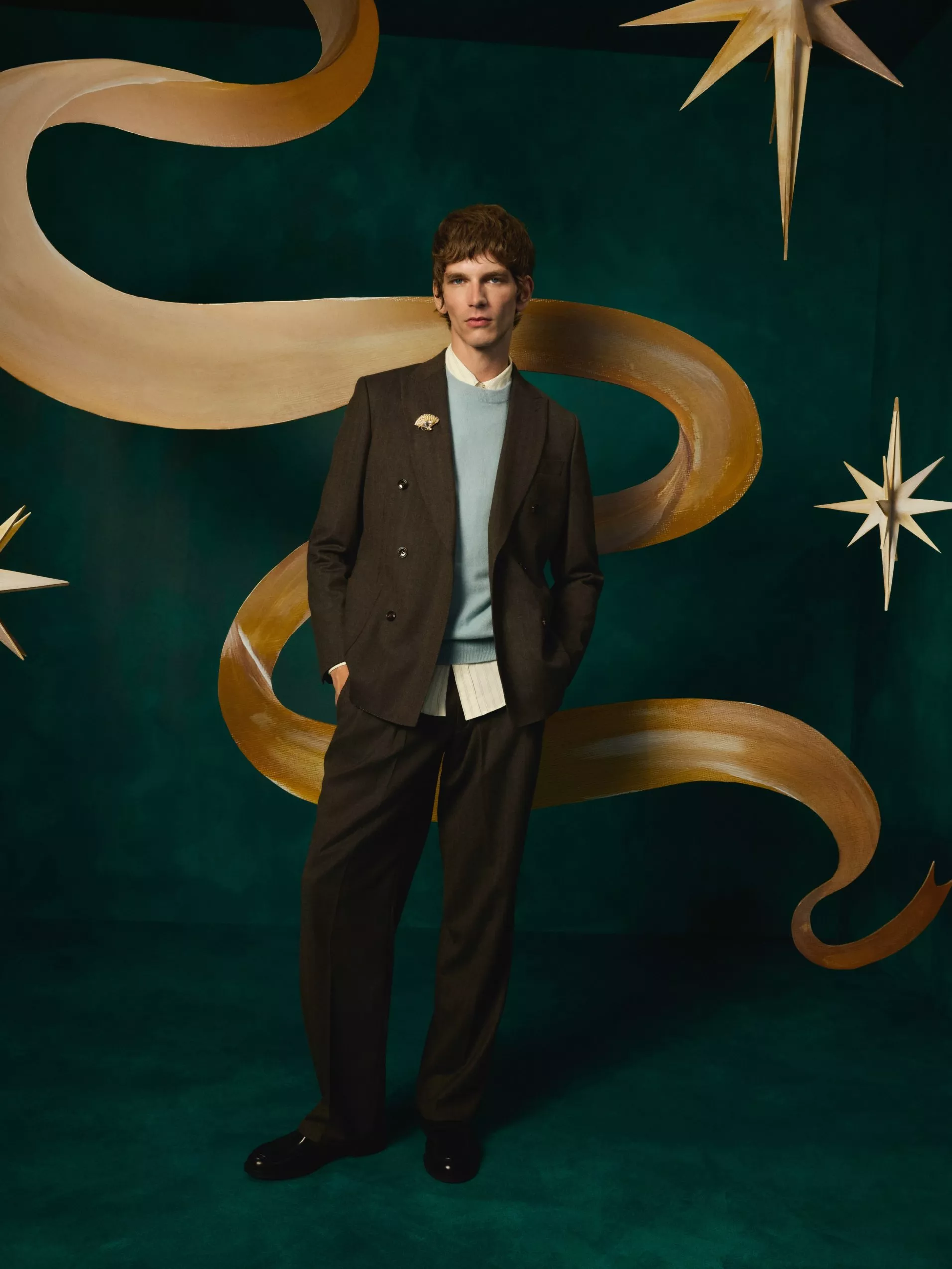 Fashion portrait of man in brown suit and blue jumper, with gold ribbon and star decorations on teal backdrop