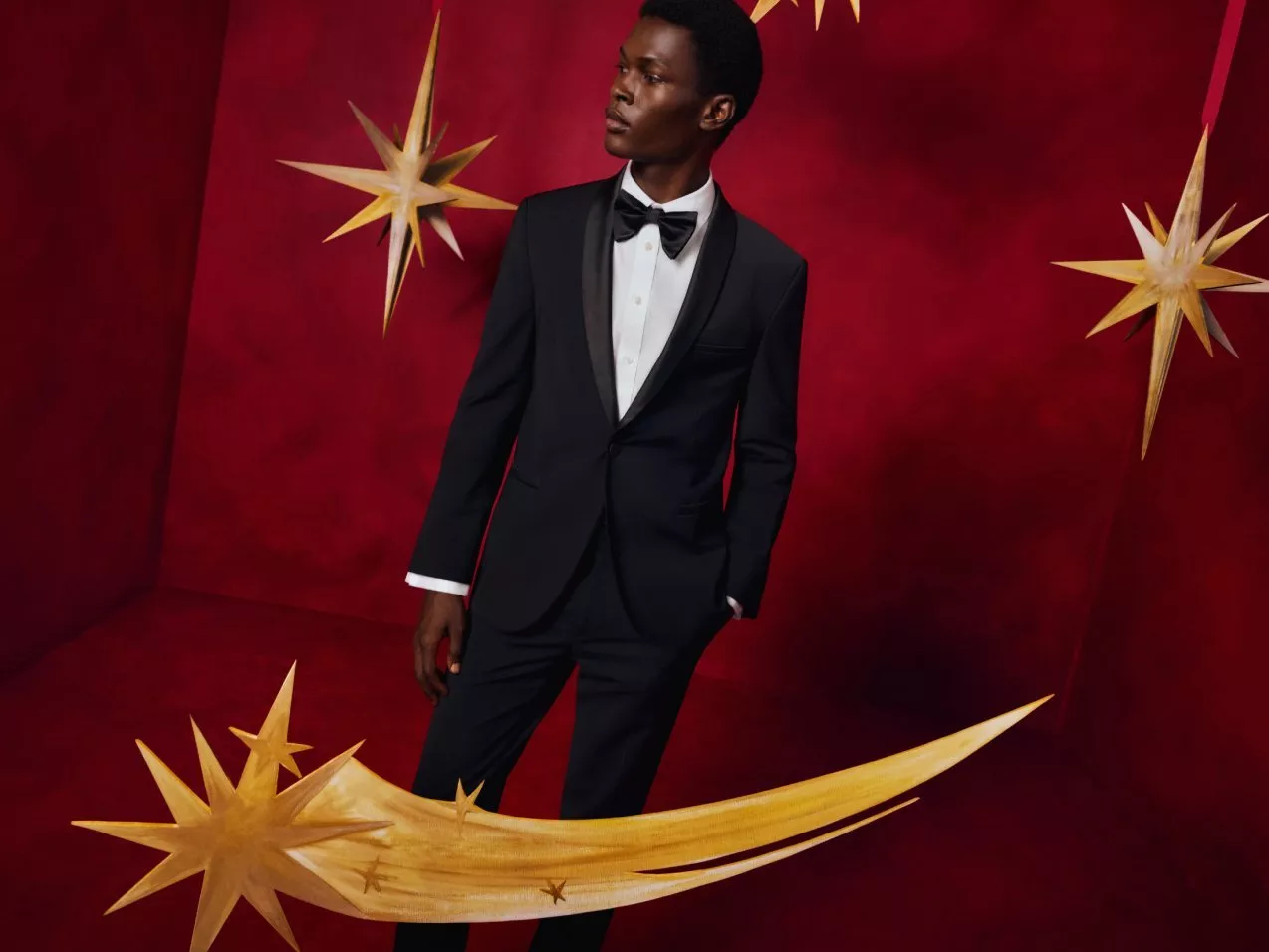 Man in black tuxedo and bow tie posing on red backdrop with gold stars, festive formalwear fashion shoot