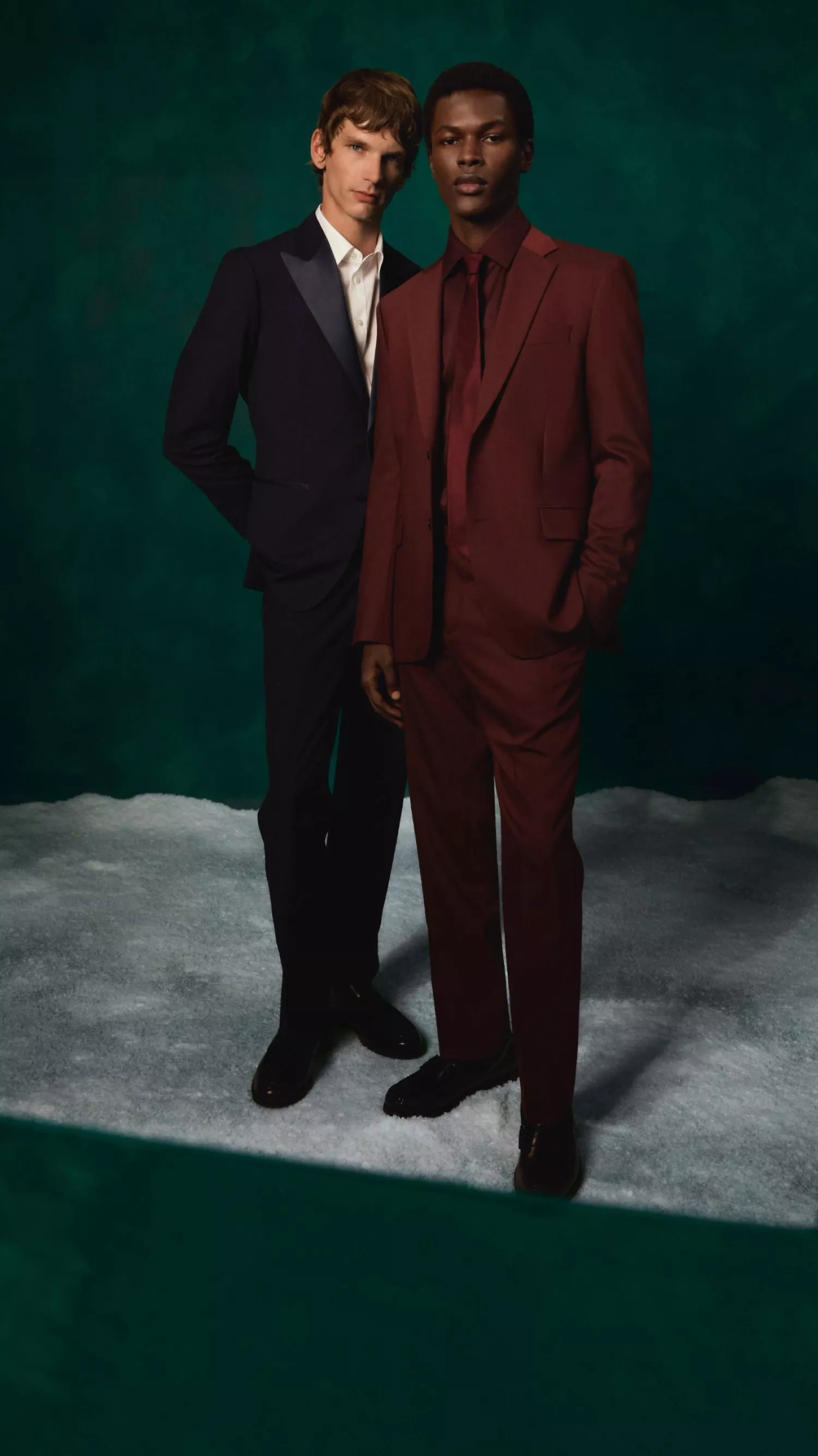 Two male models in tailored suits, one burgundy and one navy, posing in a fashion studio on green backdrop