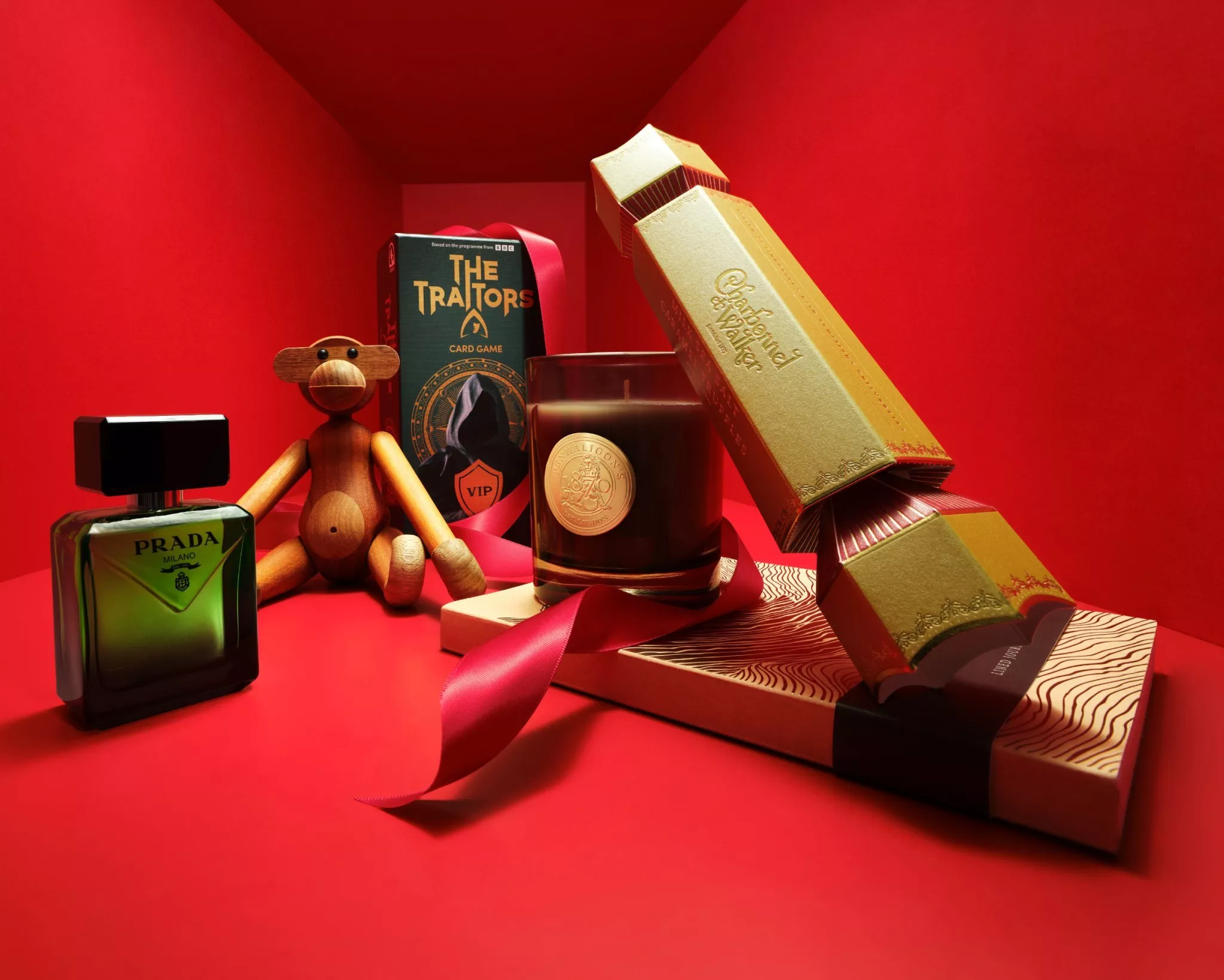 Luxury gift set on red backdrop with Prada fragrance, The Traitors card game, candle and chocolate crackers