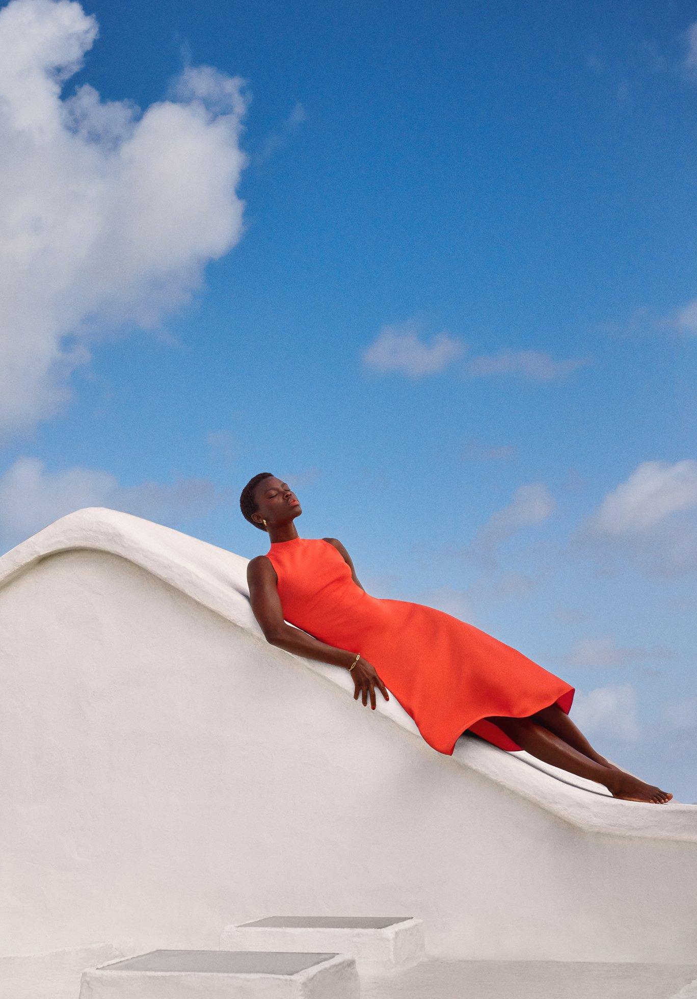 Image of a model wearing a bright orange dress, lying down on a white structure with blue sky